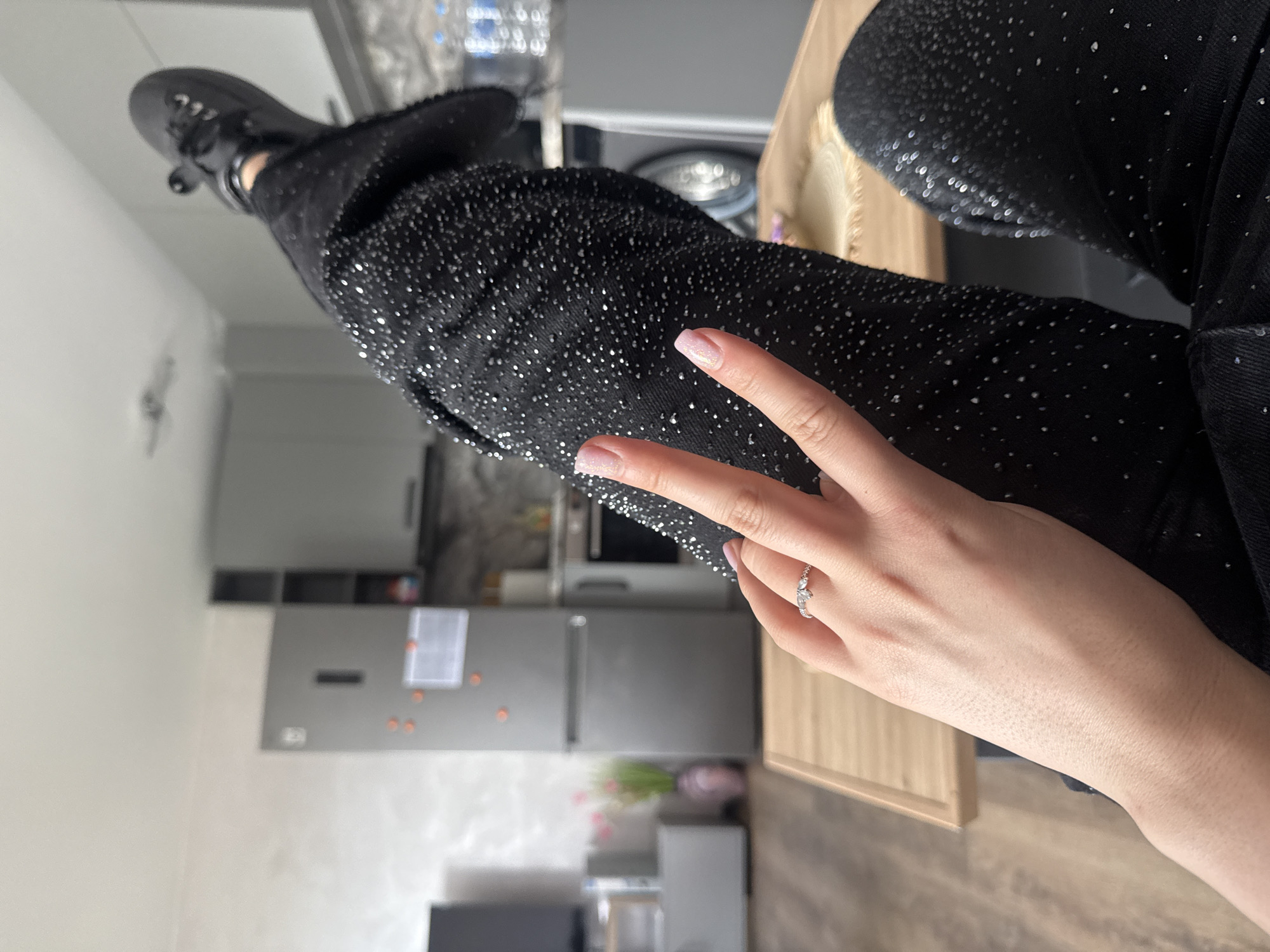 The image shows a person posing with their legs crossed and making a peace sign with their hand. They are wearing black jeans with sparkly details and black shoes. A silver ring is visible on their finger. The background shows a kitchen setting with gray cabinets, a refrigerator, a microwave, and various kitchen items. The location is indoors. Specific details regarding the location being Plovdiv, Bulgaria are not visible from the image.
