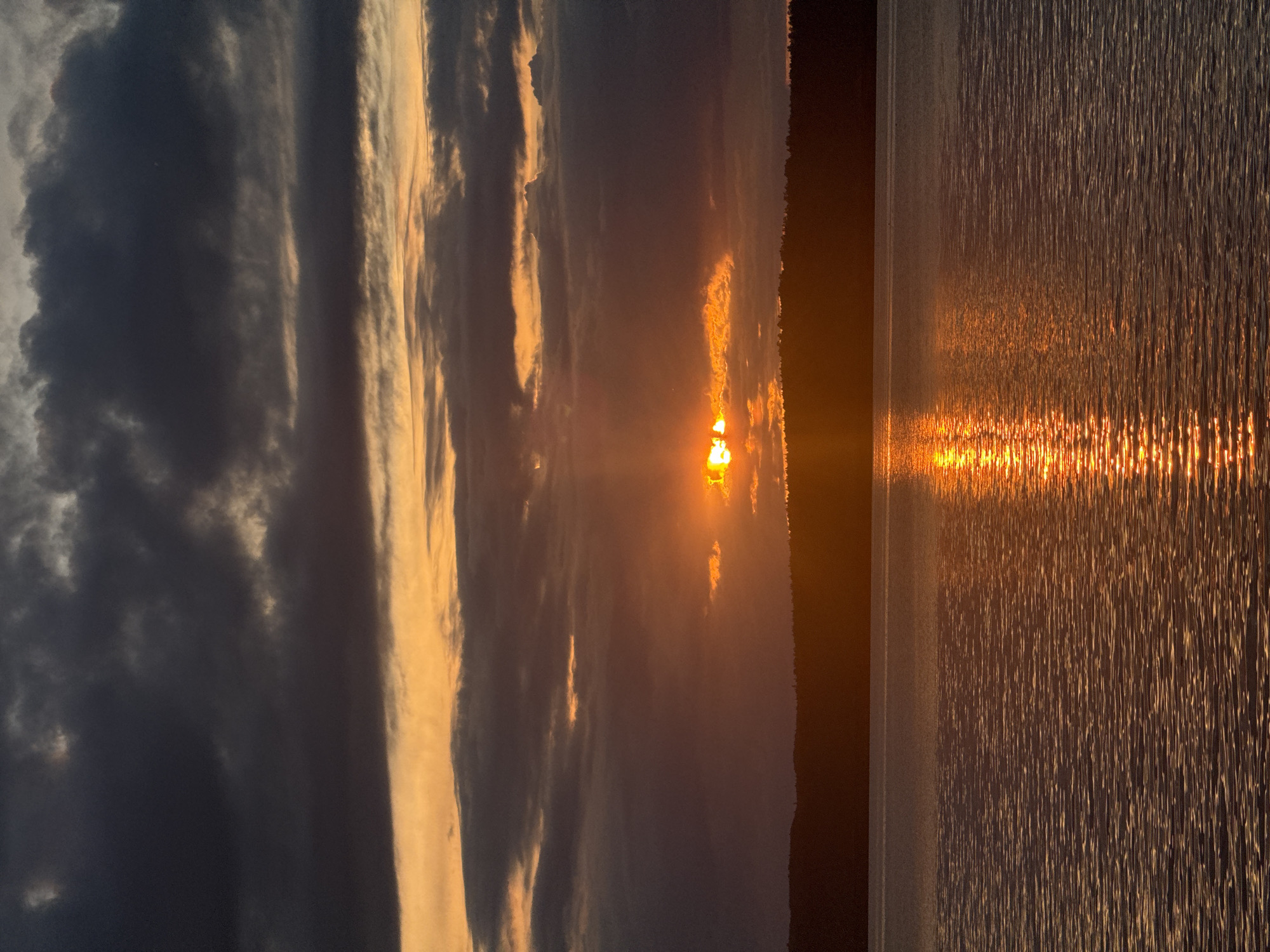 The image shows a sunset over a body of water, likely the Kama River near Chaikovsky, Russia. The sun is partially obscured by clouds, casting an orange glow across the sky and reflecting in the water. The sky features layered clouds ranging from dark grey to orange. On the horizon, a dark band of land or trees is visible. The water surface shows ripples and reflections of the sunset.