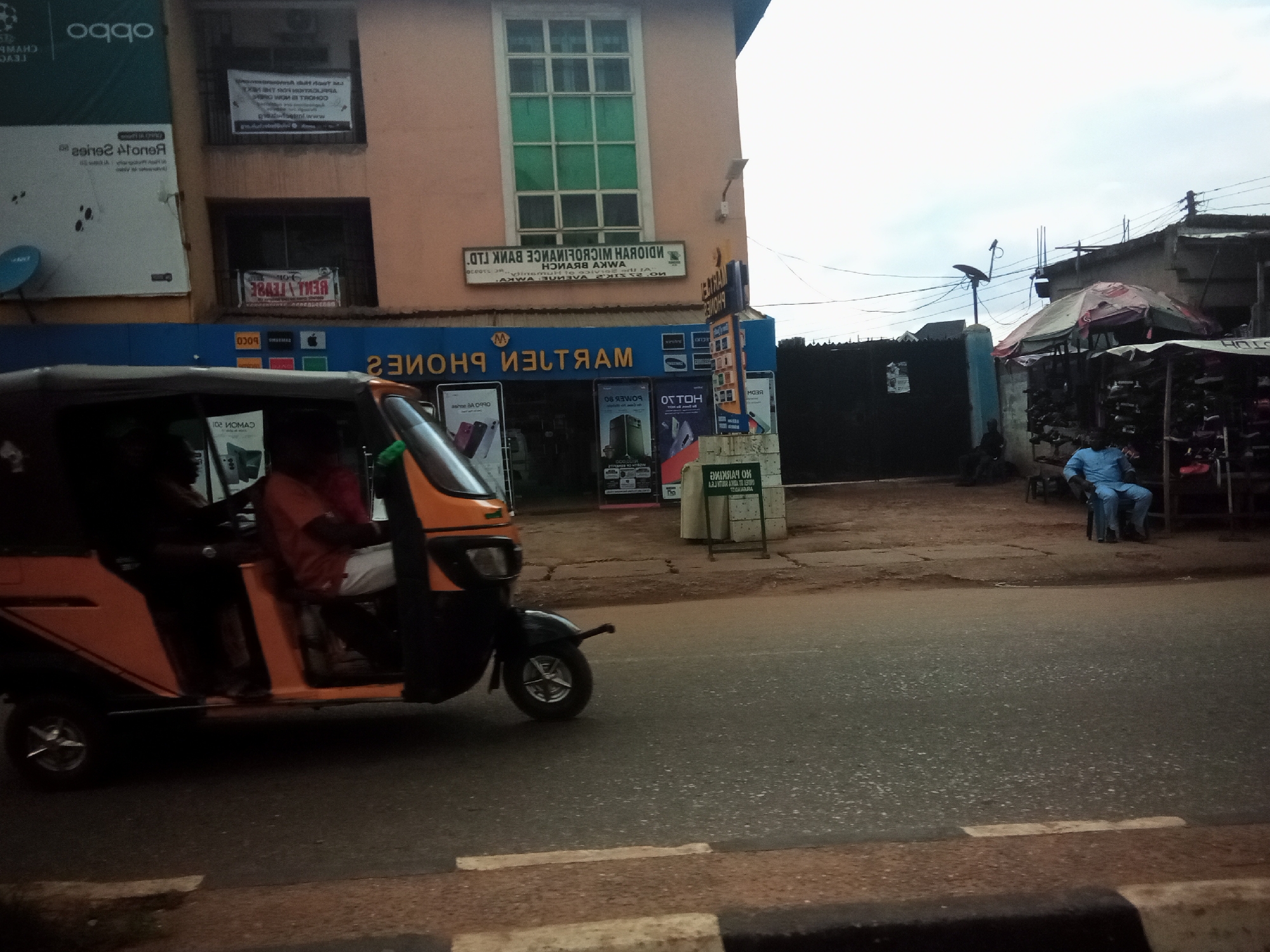 A bright orange auto-rickshaw is partially visible on the left side of the frame, in motion on a paved road. The auto-rickshaw has its doors open, revealing a person in a red shirt seated inside. Behind the auto-rickshaw, a multi-story building with a pink facade dominates the background. The building features several windows, some with green tinted glass, and a sign with text in a foreign language. A blue and yellow sign for "MARTEN PHONES" is mounted on the building's lower level. To the right of the building, a narrow street with a covered market stall is visible. A man in a light blue traditional outfit is seated on a chair in front of the market stall. Overhead, electrical wires crisscross against a cloudy sky. The scene is likely in Nibo, Nigeria, given the presence of the auto-rickshaw, the architecture, and the signage which appears to be in a local script.