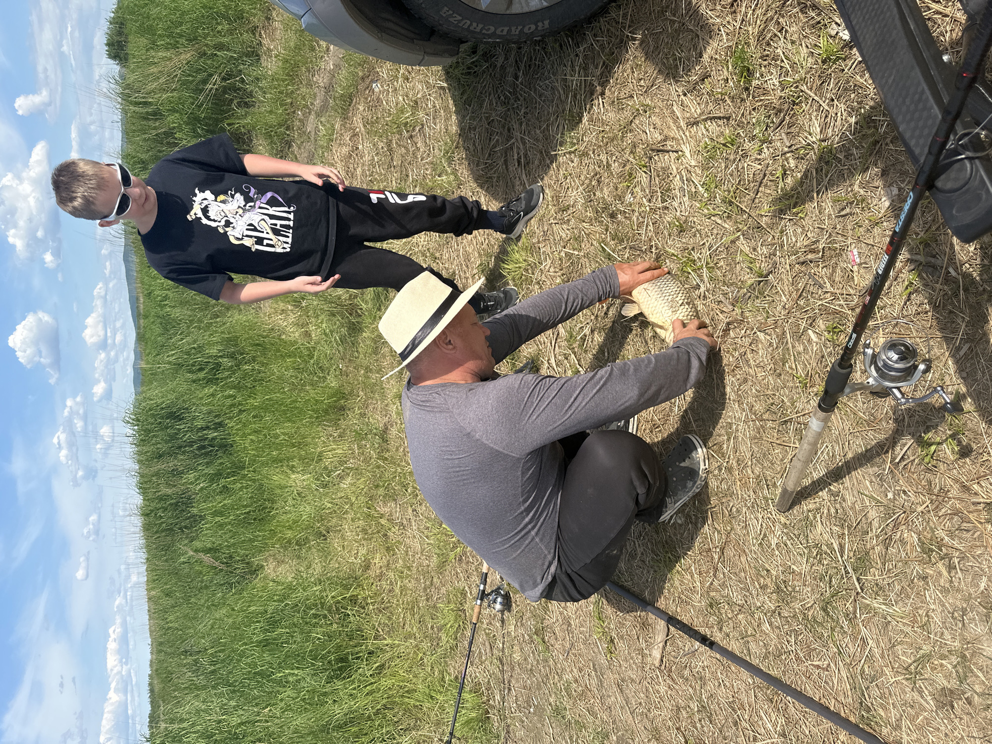 Two individuals, an adult male and a young male, are engaged in fishing in a grassy field under a partly cloudy sky. The adult male, wearing a straw hat and a grey long-sleeved shirt, is crouched down, holding a caught fish. The young male, wearing a black t-shirt and sunglasses, stands behind him, observing. Several fishing rods are visible, resting on the ground or in the foreground. A vehicle is partially visible on the right side of the frame. The scene appears to be outdoors in a natural setting, possibly near a body of water, given the fishing activity. The location is identified as Im. Tel'mana, Russia.