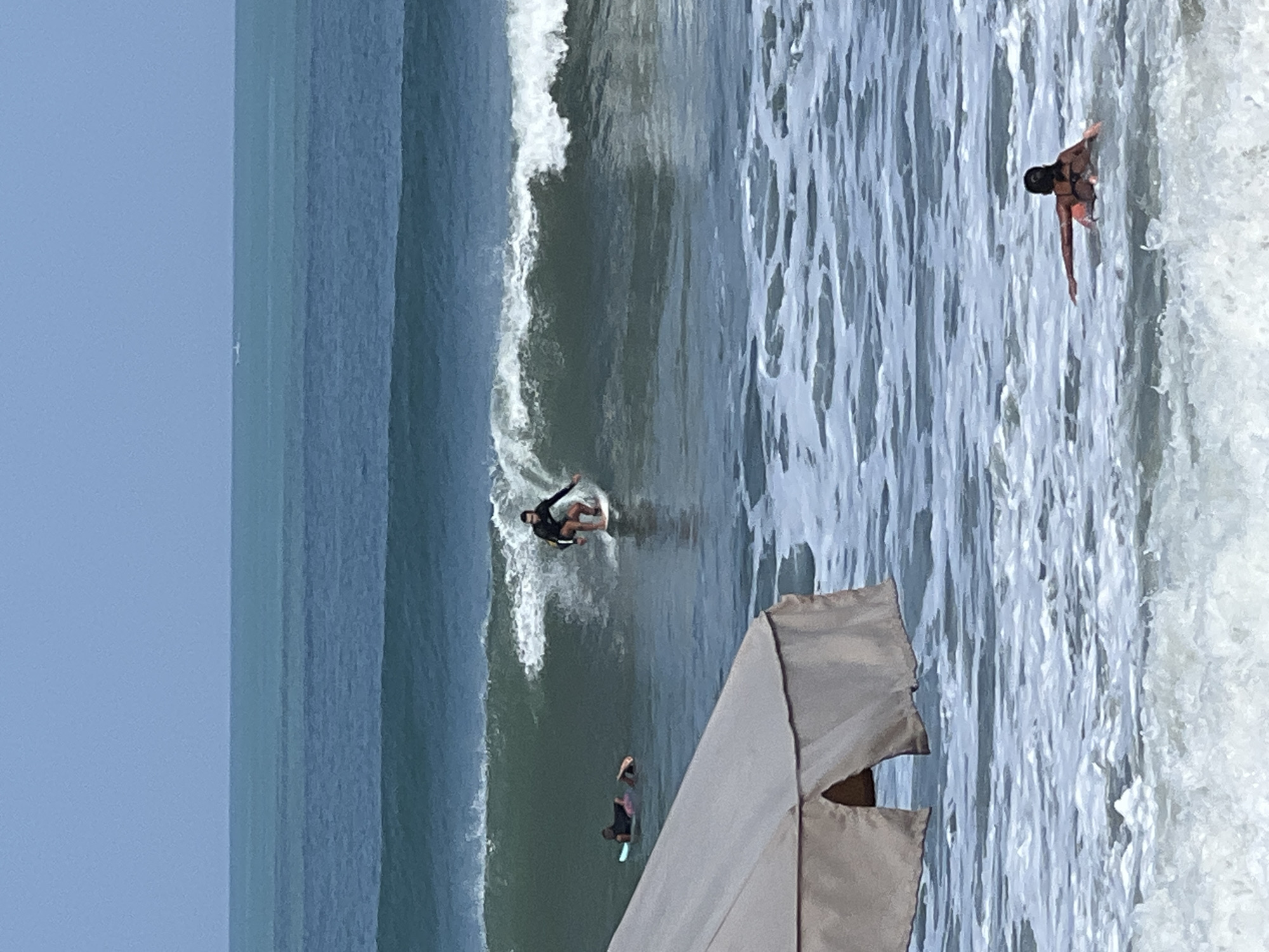 In Kuta, Indonesia, a surfer in a black wetsuit rides a wave, appearing mid-action on a white surfboard. Behind him, two individuals are on surfboards in the water, with one lying down. In the foreground, the edge of a large, tan-colored canopy or umbrella is visible, partially obscuring the view of the ocean. A person wearing a black bikini top and orange bottoms is in the water in the lower right corner, facing away from the camera. The sea is a deep blue-green, with white foam and waves indicating active conditions. The sky above is a clear, light blue.