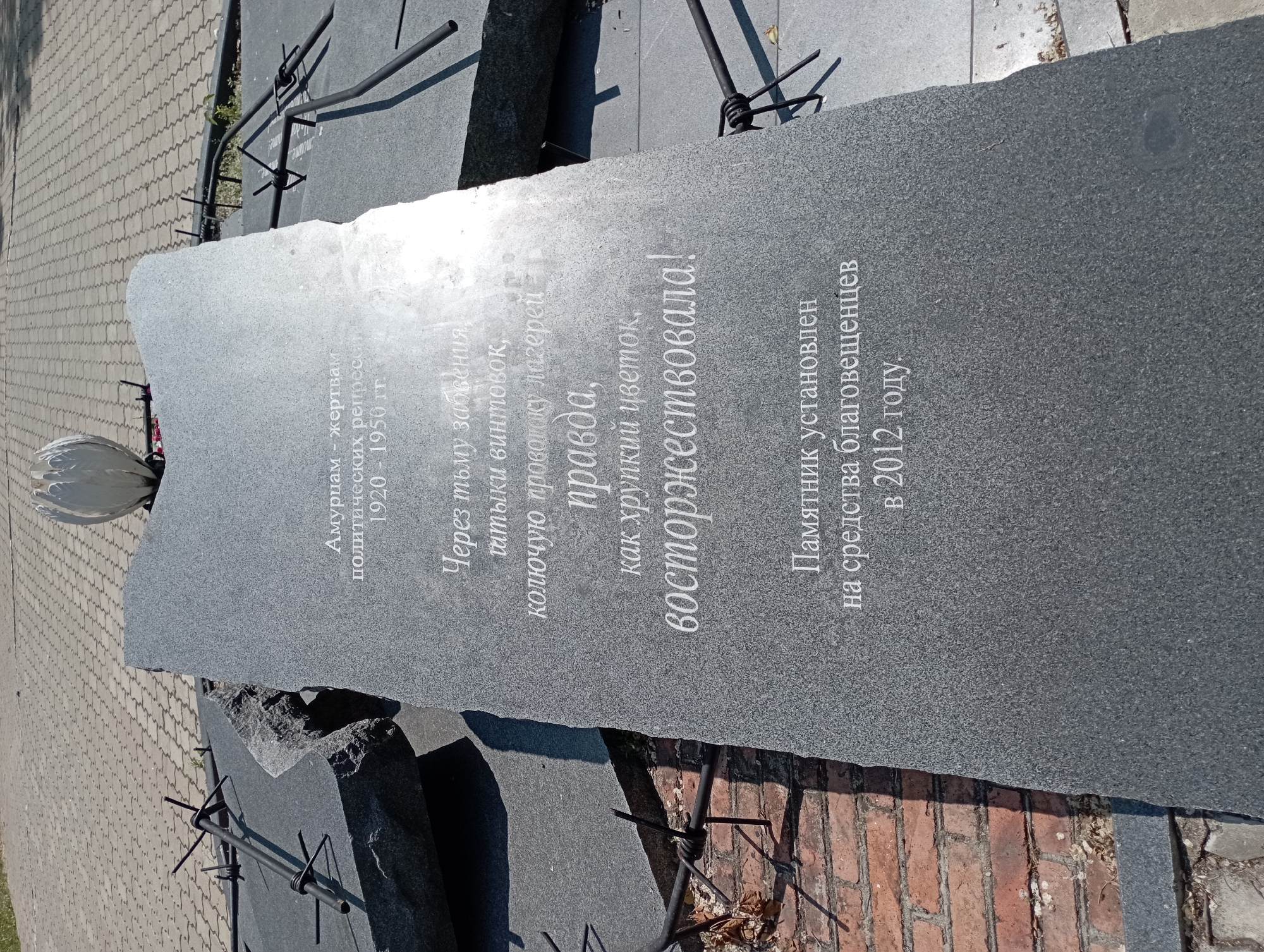 This image depicts a monument in Blagoveshchensk, Russia, dedicated to victims of political repression between 1920 and 1950. The monument is a large, upright slab of dark grey granite, with an uneven, broken top edge. Inscribed on its surface in white lettering are the words: "Амурцам - жертвам политических репрессий 1920 - 1950 гг. Через тьму забвения, пытки винтовок, колючую проволоку лагерей, правда, как хрупкий цветок, восторжествовала! Памятник установлен на средства благовещенцев в 2012 году." This translates to: "To the people of Amur - victims of political repression 1920 - 1950. Through the darkness of oblivion, the torture of rifles, the barbed wire of camps, truth, like a fragile flower, triumphed! The monument was erected with funds from the citizens of Blagoveshchensk in 2012."
The monument is surrounded by other dark granite slabs, some of which appear broken or dislodged, creating a fragmented landscape. Barbed wire, twisted and sharp, is prominently displayed, woven around metal poles that emerge from the ground and the granite slabs, symbolizing the harsh realities of repression. In the background, a paved walkway with a geometric pattern is visible, along with a bright, stylized white sculpture resembling a blooming flower or a lotus. The scene is set outdoors under daylight, with shadows indicating a sunny day. The lower left corner shows a brick surface, suggesting the proximity to a building or a different ground level. The overall impression is somber and reflective, with the elements of the monument and its surroundings conveying themes of suffering, resilience, and ultimate triumph of truth.