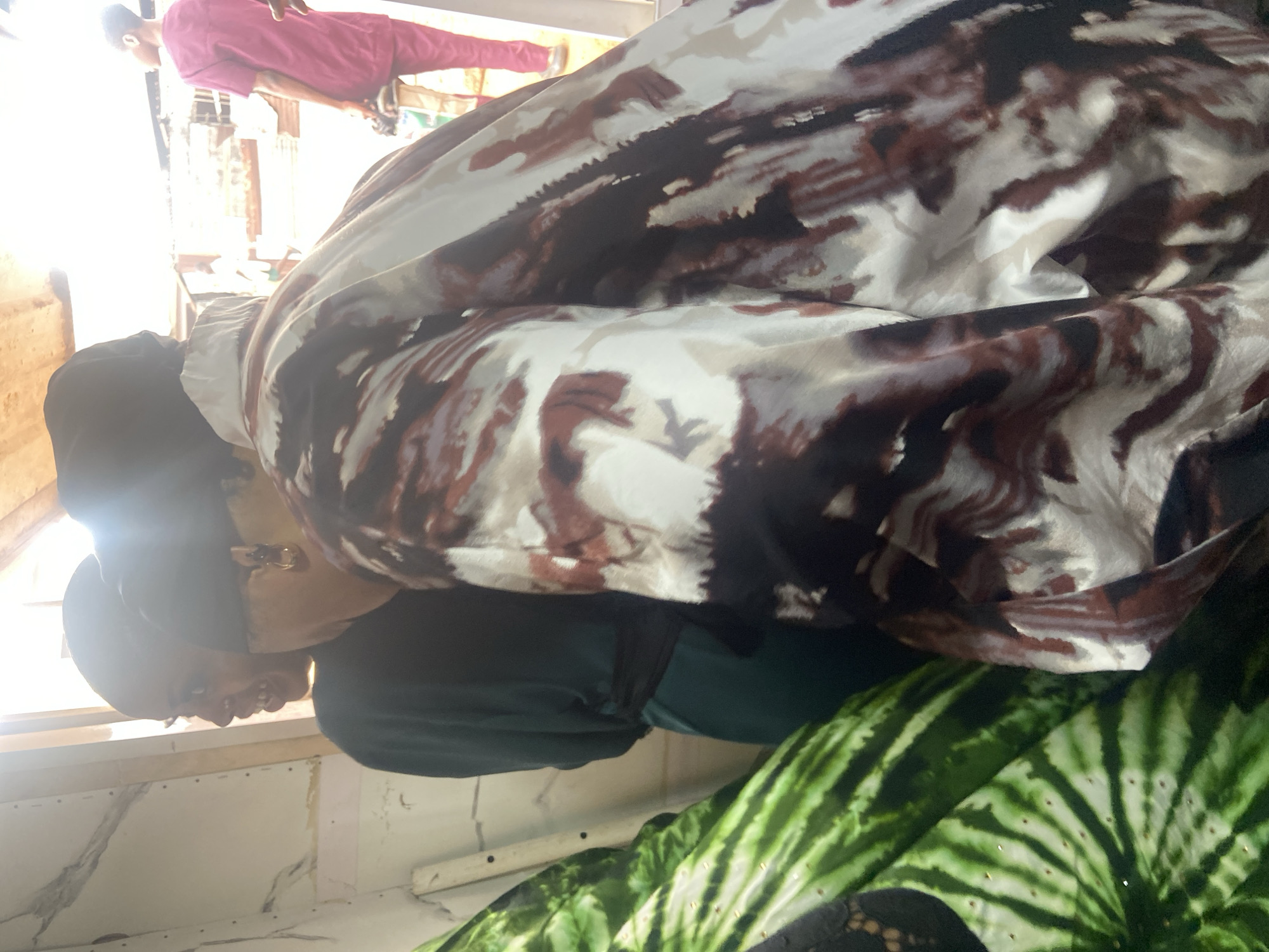 The image, apparently taken inside a building in Okengwe-Obehira, Nigeria, features a woman wearing glasses, a black cap, and a green top. She is positioned near a window, which is illuminating her face. Part of a patterned fabric drapes over her, obscuring much of her body. In the background, another person can be seen wearing pink clothing. The environment appears to be a simple interior with a white wall.