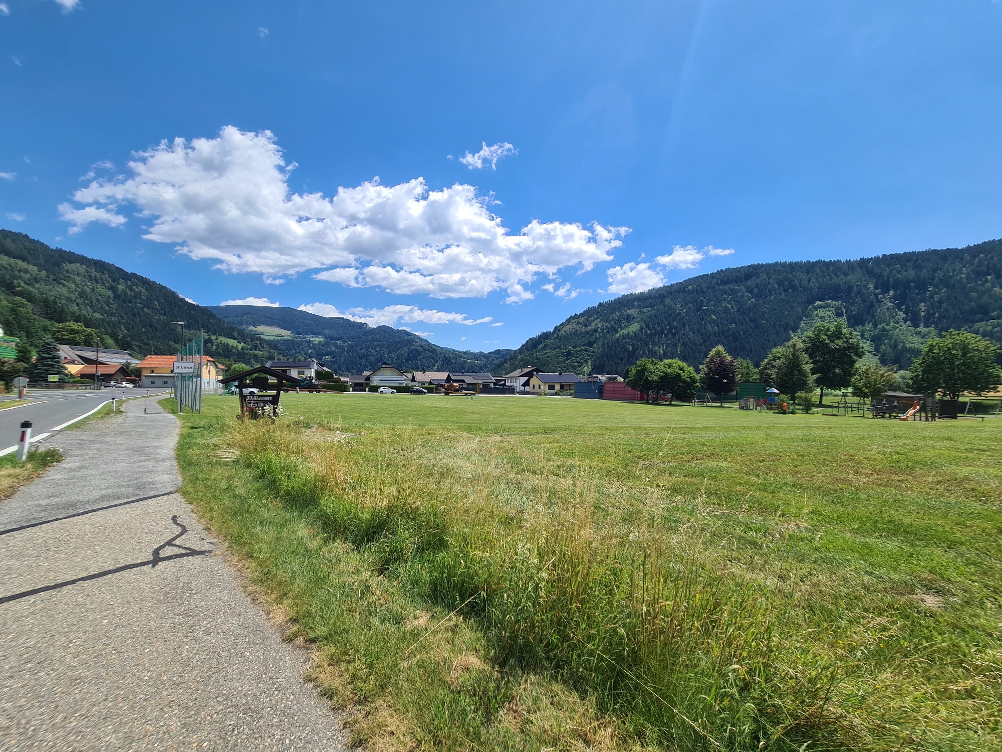 The image captures a bright, sunny day in Feldkirchen in Kärnten, Austria, focusing on a grassy field adjacent to a paved pathway. The field extends towards a cluster of buildings and is bordered by rolling, tree-covered hills under a clear blue sky with scattered white clouds. A road is visible on the left, with a white line marking its edge and a few cars parked in the distance. On the field, a playground area with swings, a slide, and other equipment can be observed. A wooden structure with a sign reading "Willkommen" (Welcome) stands near the pathway. The foreground is dominated by tall, green grass with some dry, brown patches. The overall scene depicts a tranquil, semi-rural environment.