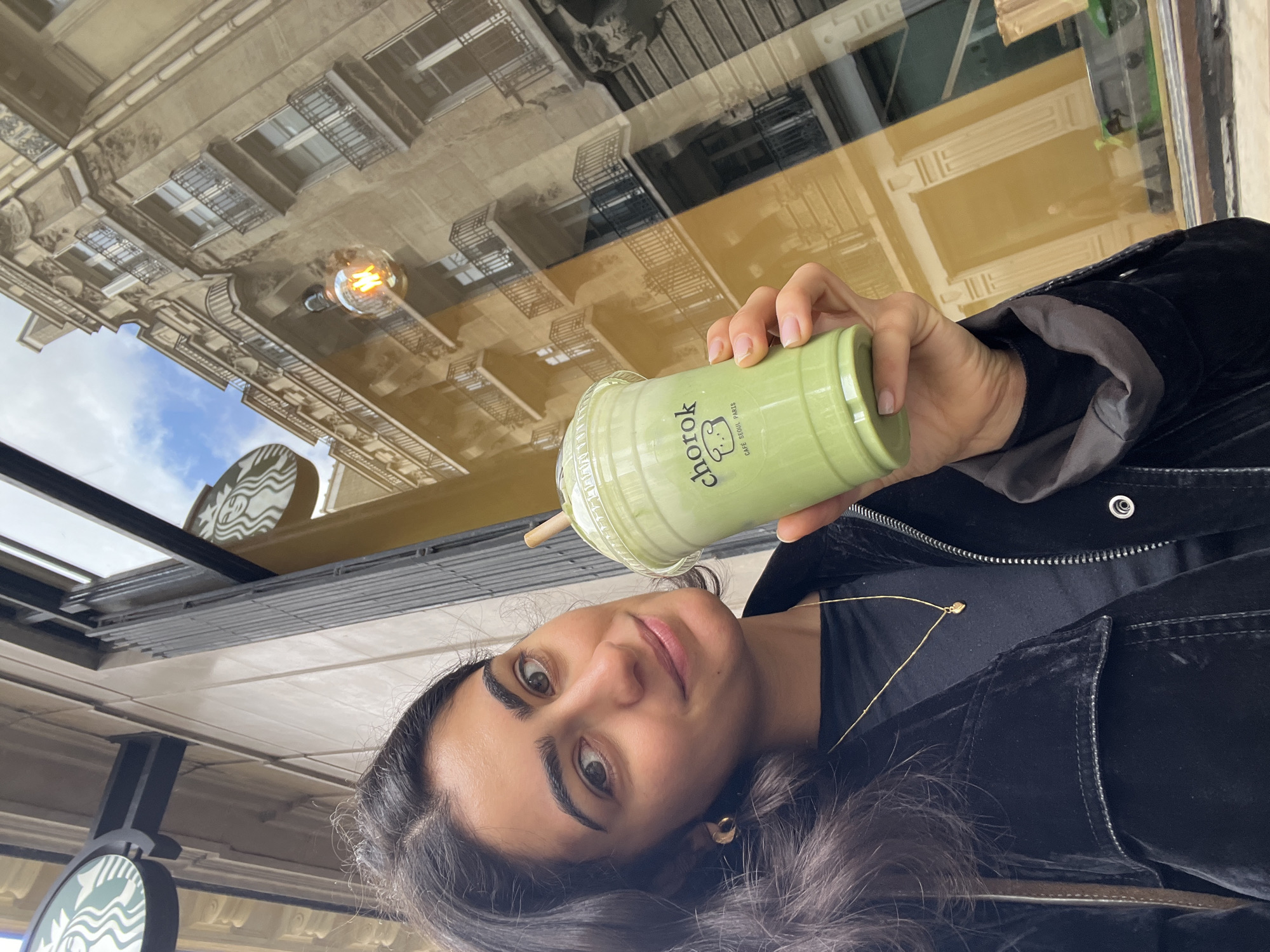 A person is holding a "chorok" labeled green drink in front of a window. The window reflects the architecture of a Parisian building and a hanging light fixture. The person has dark hair and is wearing a black jacket and a gold necklace. The image was taken inside a cafe.