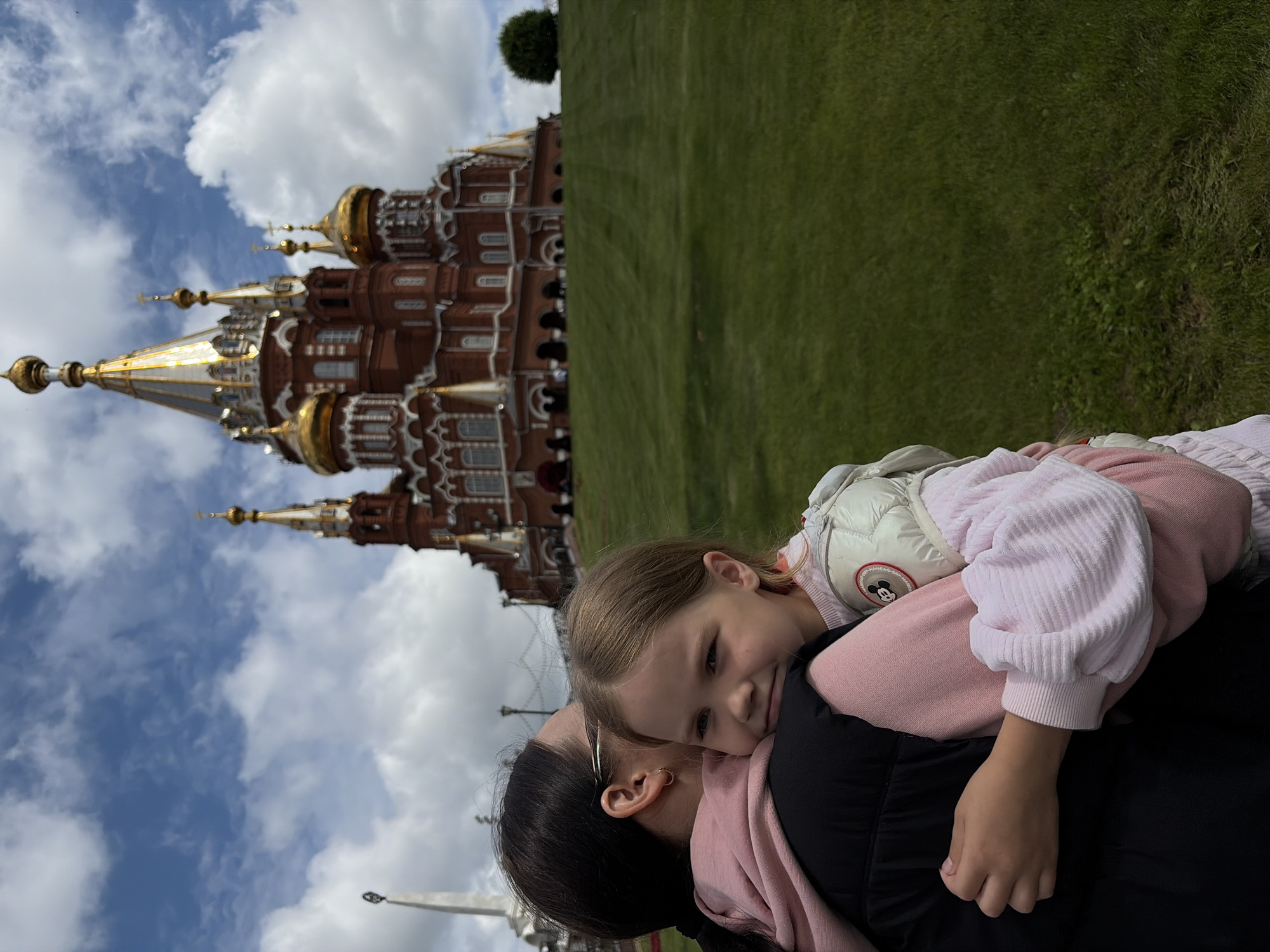 This media file shows a close-up of a young girl, with blonde hair, resting her head on the shoulder of an adult, who has dark hair. The girl is wearing a pink and white jacket with a cartoon character patch and a pink sweater. The adult is wearing a black jacket and a pink scarf. They are outdoors, with a large, ornate red brick building with golden domes in the background, suggesting a significant historical or religious site. A grassy lawn is visible to the right of the building. The sky is cloudy and blue, indicating daytime. The image conveys a feeling of affection and closeness between the two subjects. The location is identified as Izhevsk, Russia. No visible text is present in the image.