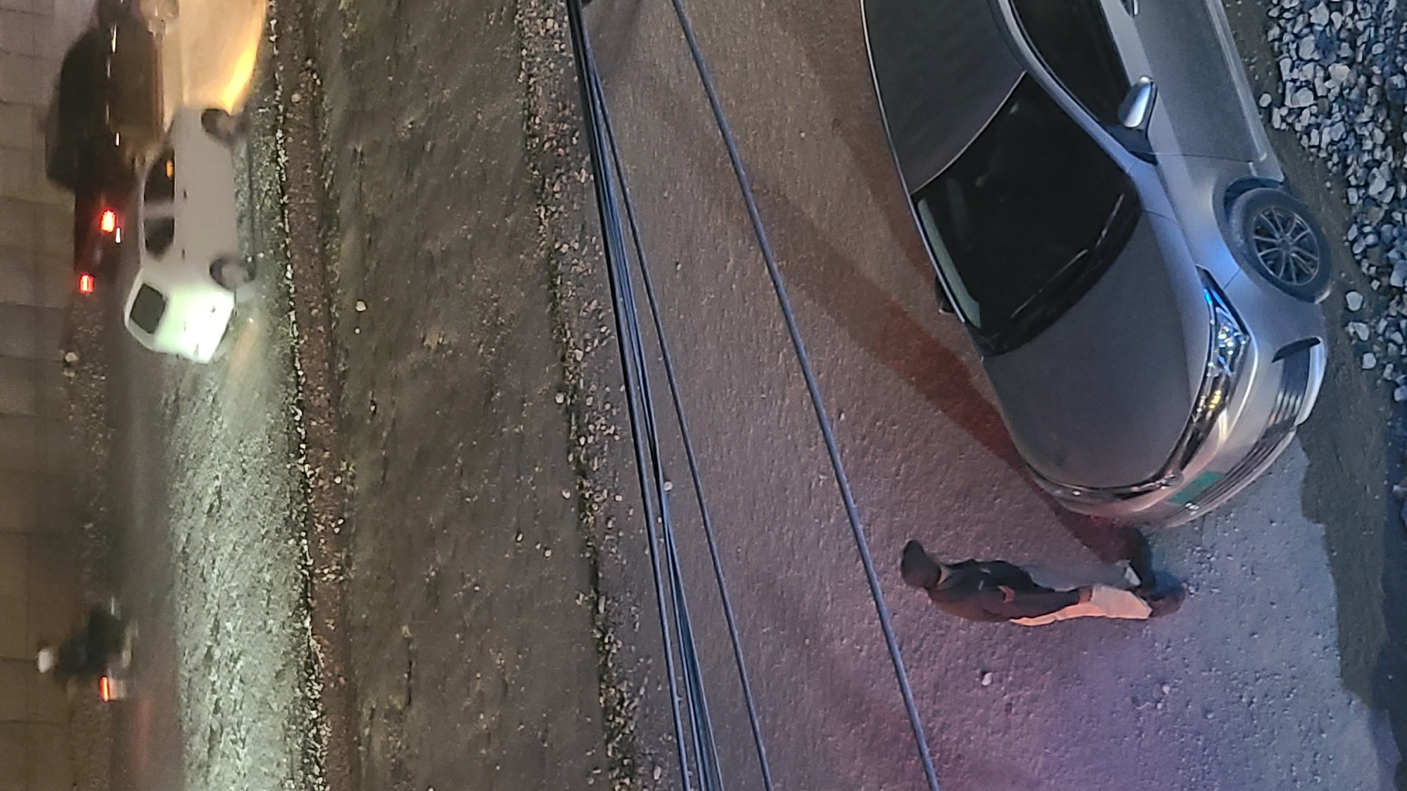 The image, taken from an elevated perspective, shows a street scene in Quetta City Tehsil, Pakistan. A silver car is parked on the right side of the frame. A person wearing dark clothing with light-colored pants stands near the front of the car. Power lines run diagonally across the center of the image. On the left side of the frame, there is a white vehicle, followed by a motorcycle. The environment includes gravel and paved areas.