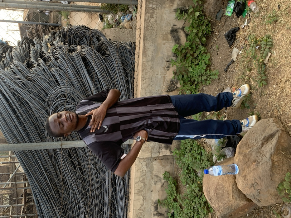 A man wearing a striped shirt and blue pants poses next to a large pile of metal rebar. He has his right hand on his chest and is wearing a watch. Behind him, there is a concrete wall and overgrown foliage. He is standing on the ground, where there is some trash scattered around. A water bottle is placed on a rock. The man appears to be the focal point of the image, with the rebar pile as a secondary element of the background.