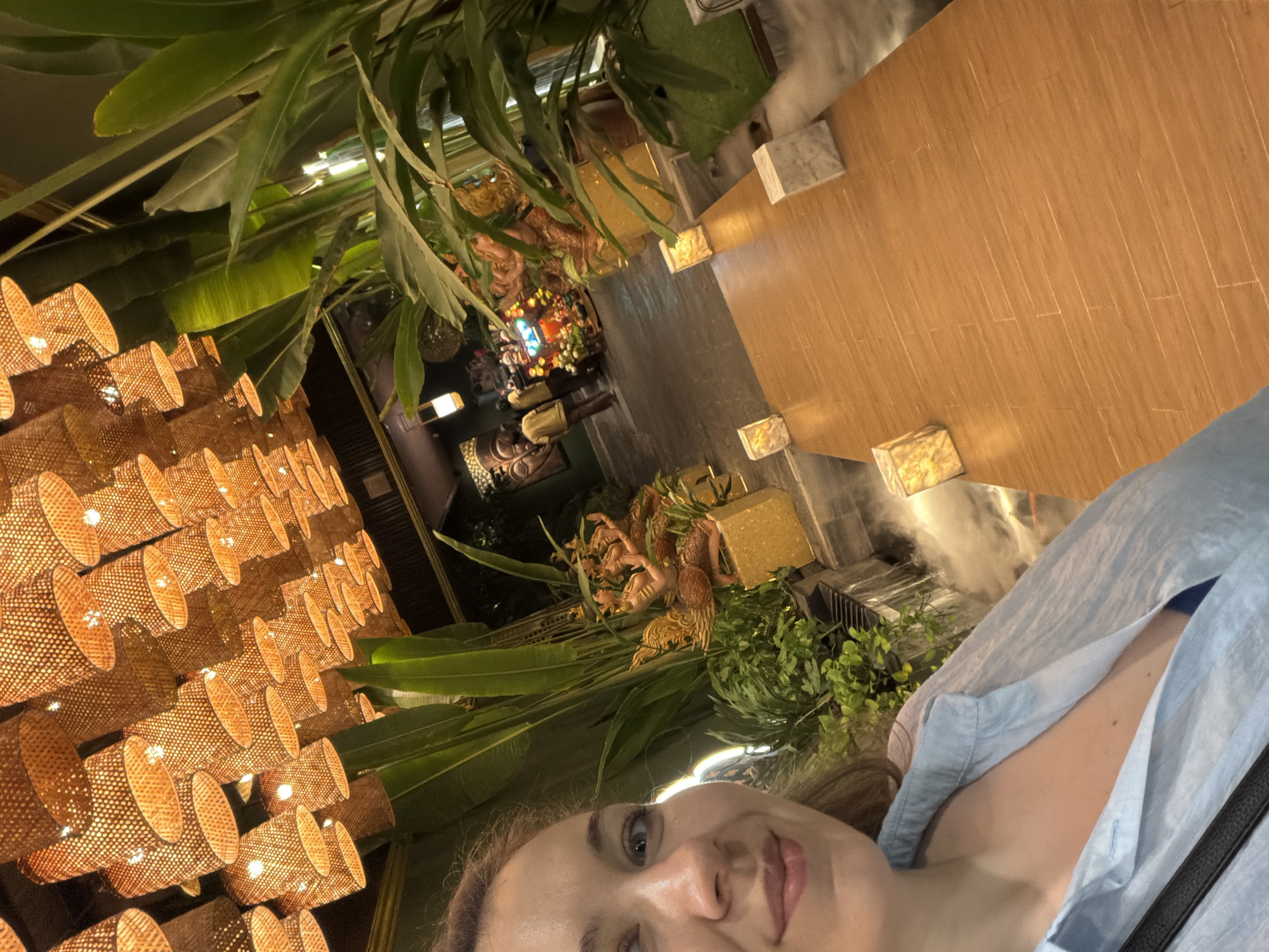 This rotated selfie captures a woman in a light blue collared shirt, looking directly at the camera, with a neutral to slight smile. The setting is an elaborately decorated indoor or covered outdoor space, likely in Nha Trang Ward, Vietnam, given the cultural cues.
The background is rich with decorative elements, suggesting an evening or nighttime ambiance due to the prominent artificial lighting. To the left, a wall or ceiling is adorned with numerous warm-toned, woven basket-like light fixtures, casting a soft, inviting glow. Lush green tropical foliage, including large banana-like leaves and smaller plants, is integrated throughout the decor.
In the mid-ground, amidst the greenery, are several ornate, golden-hued sculptures, appearing to depict mythical figures or deities. A dark-colored Buddha bust or statue is also visible, adding to the Southeast Asian aesthetic. The right side of the image features a smooth, light brown wooden surface, possibly a floor or large table, with decorative square blocks (perhaps marble) placed at intervals. A delicate mist or smoke is visible near the bottom right, possibly from a humidifier or a decorative water feature. The overall scene evokes a serene, exotic, and well-designed environment, typical of a resort, restaurant, or spa. No visible text is present in the image.
