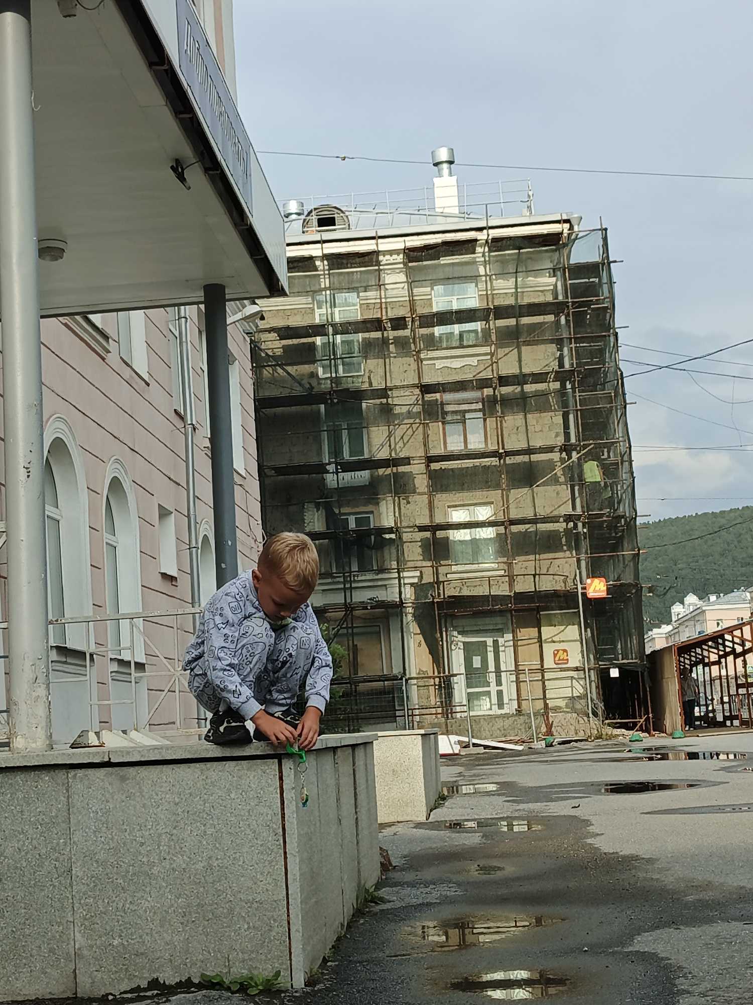 A young boy wearing a light blue pajama set with a cartoon pattern is crouched on a concrete ledge. He is looking down and playing with a green object, possibly a toy or a keychain, held in his hands. The boy is positioned in front of a light pink building with arched windows. Behind this building, a taller, older building is covered in scaffolding and netting, suggesting it is undergoing renovation or construction. The sky is overcast. The scene appears to be in an urban setting, with a wet asphalt ground indicating recent rain, featuring several puddles reflecting the surroundings. The overall impression is that of an outdoor street scene in Zlatoust, Russia.