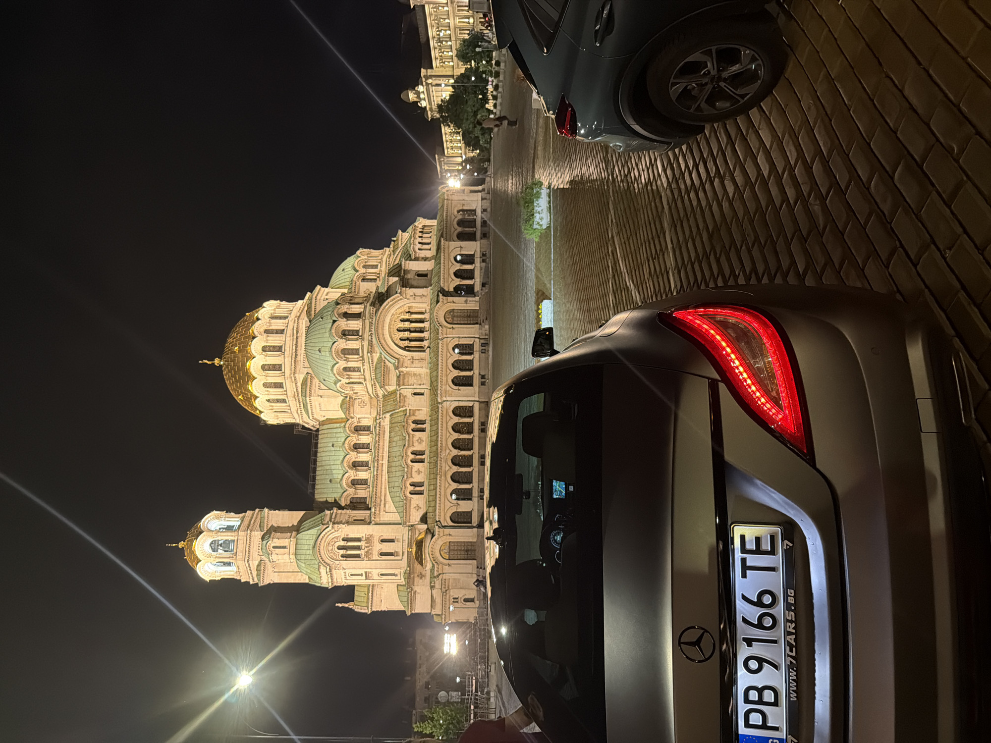 The image presents a nighttime urban scene centered around a large, ornately designed cathedral-like building with multiple domes, some golden and others green. The building is brightly illuminated, highlighting its intricate architectural details, arches, and windows against a dark night sky.
In the foreground, dominating the right side of the frame, is the rear of a dark grey or matte silver car, possibly a Mercedes-Benz given the visible logo on the trunk. Its red taillight is lit, and it bears a European-style license plate with the registration "PB 9166 TE". Below the numbers, the web address "www.7CARS.bg" is clearly visible, suggesting a car dealership or rental service. The car is parked on a textured paved surface, which appears to be made of cobblestones or bricks.
Behind the prominent car, another dark car is partially visible. The paved area extends into the mid-ground, leading towards the grand building. Streetlights emit strong light, creating visible lens flares. In the distance, beyond the immediate street level, other buildings can be faintly seen. At least one person is discernible walking in the background near the base of the main illuminated structure. The scene portrays an active urban environment at night, with artificial lighting accentuating the architectural grandeur.