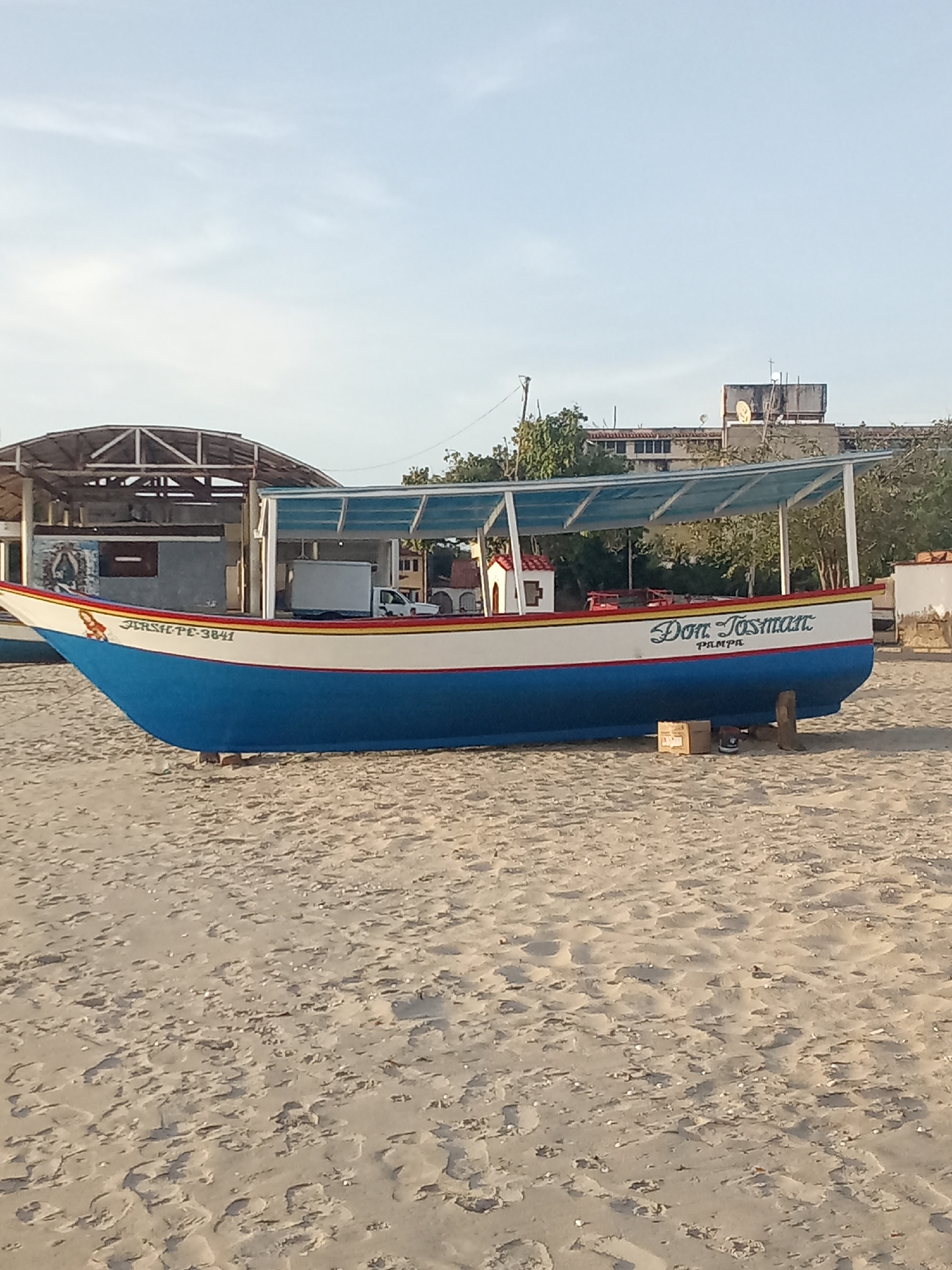 The image depicts a blue and white fishing boat, named "Don Josman PAMPA," resting on a sandy beach. The boat has a covered seating area. In the background, there is a building with a partially visible facade and a taller building with a satellite dish on its roof. The scene appears to be located in Juan Griego, Venezuela. The sky is mostly clear. The beach is sandy and has an uneven texture. There are no people visible in the immediate foreground. The boat is elevated off the ground, supported by wooden planks and a cardboard box. The other boat is also standing in the sand.