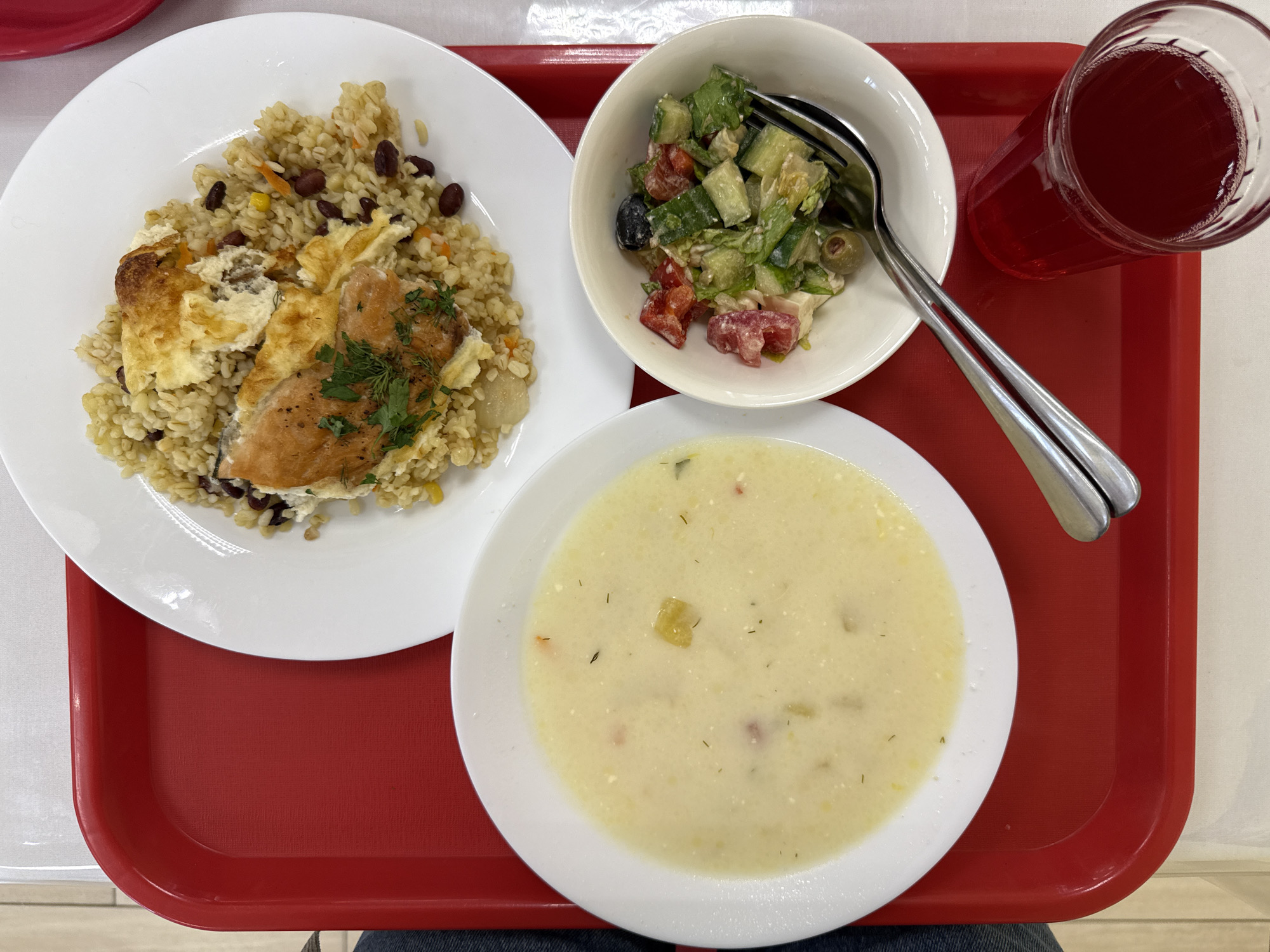 An overhead, slightly angled shot captures a red cafeteria tray on a white surface. The tray holds a meal consisting of a white bowl of creamy soup with visible diced vegetables, a white plate with a portion of rice pilaf topped with a piece of baked fish and a creamy sauce, and a small white bowl of salad containing chopped tomatoes, cucumbers, lettuce, olives, and feta cheese. A fork rests in the salad bowl. To the right of the tray, a clear glass filled with a dark red beverage is partially visible. The overall impression is that of a simple, prepared meal. There is no discernible location in the city of Dzerzhinsk, Russia.