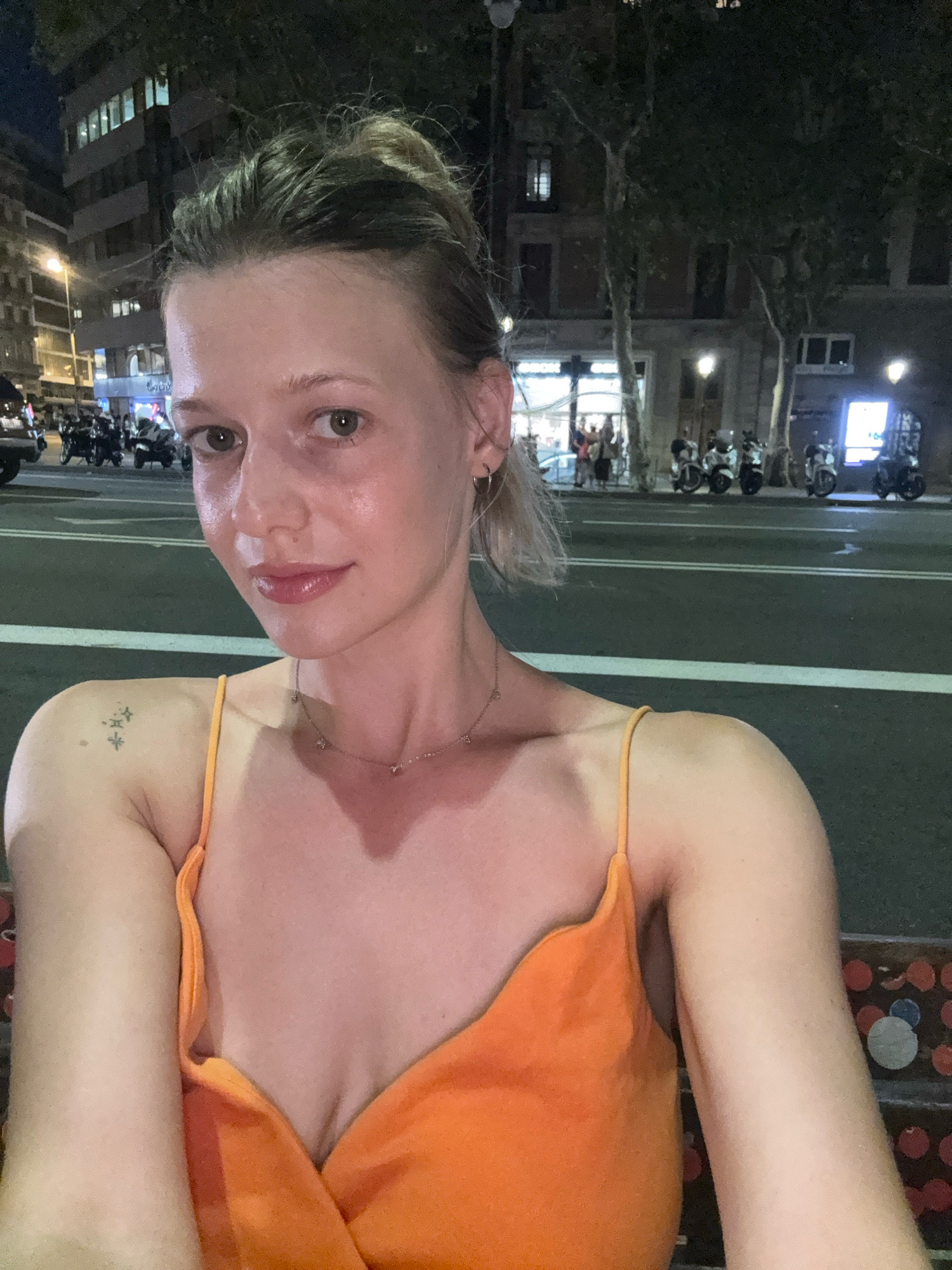 A medium shot, selfie-style image shows a woman with light skin and blonde hair pulled up in a bun. She is wearing an orange, spaghetti-strap top. Her left arm is raised, and she has a small tattoo on her left shoulder. A delicate necklace with small charms is visible around her neck. She is in Barcelona, Spain, at night. In the background, there are streetlights, buildings with illuminated windows, and several parked motorcycles. The street is paved, and white lines mark the lanes.