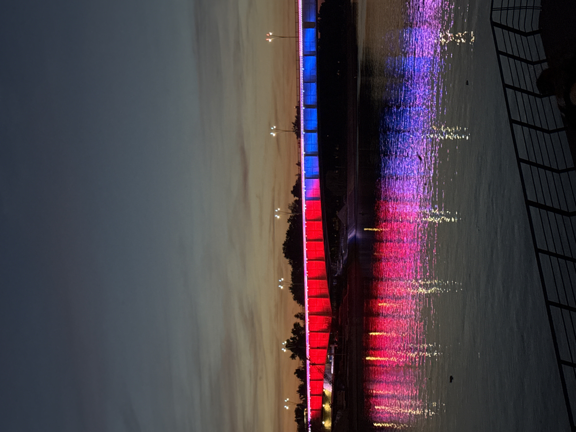 The image shows a bridge illuminated with red, white, and blue lights at night, reflected in the water below. The bridge spans a body of water, possibly a river, and its lights cast colorful patterns on the water's surface. The sky is dusk or night, with some faint color remaining. A metal railing is visible on the right side of the image. The location is likely Belgrade, Serbia, given the colors are those of the Serbian flag.