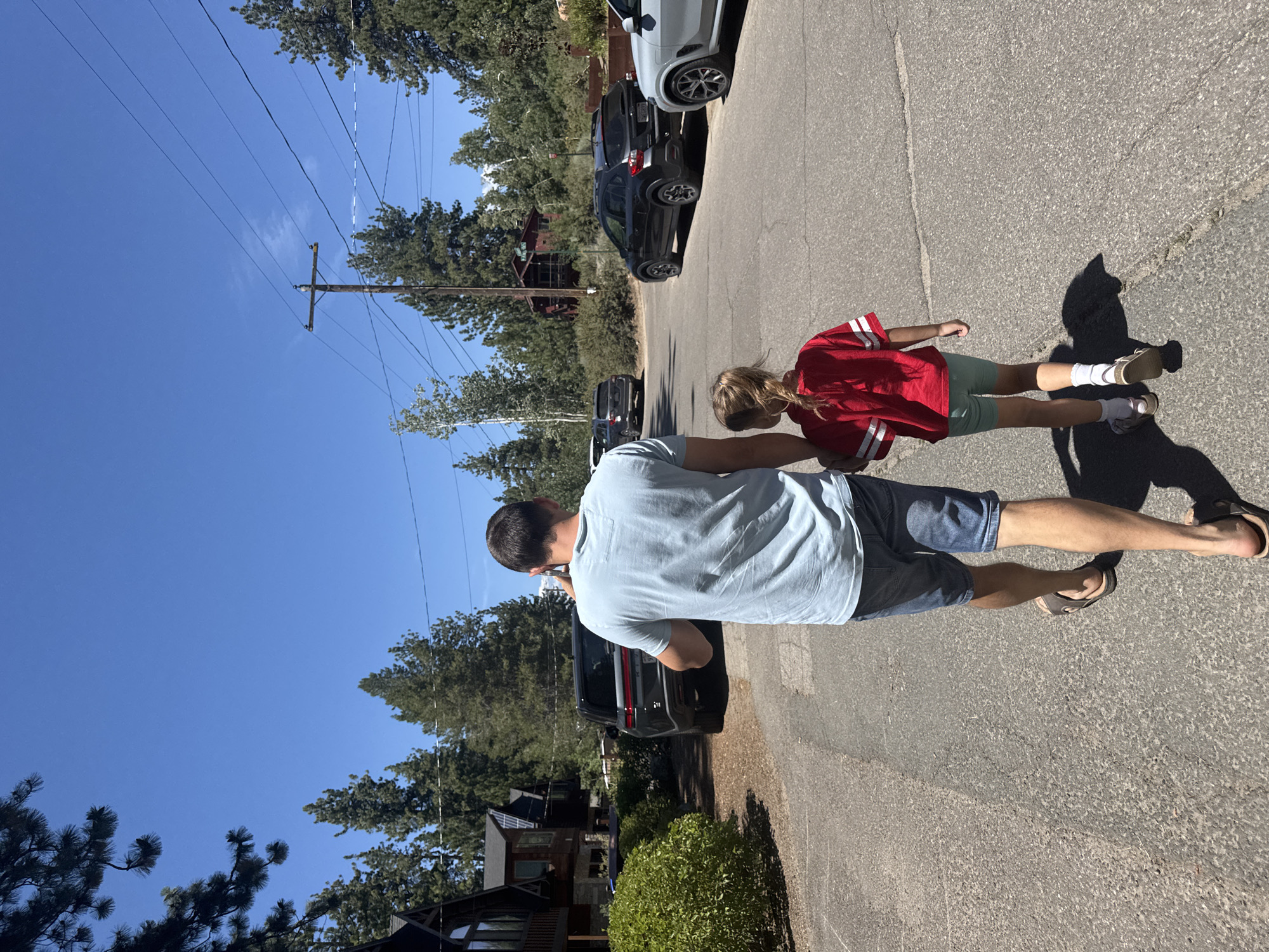 A man and a young girl walk hand-in-hand down a paved street in South Lake Tahoe, California. The man, viewed from behind, wears a light blue t-shirt and denim shorts, with brown sandals. He is holding a smartphone to his ear with his left hand. The girl, also seen from behind, wears a red t-shirt with white stripes on the sleeves and light green shorts, with white socks and sandals. They are walking away from the camera, towards a residential area with houses and parked cars visible on either side of the street. Tall pine trees and a bright blue sky form the backdrop, suggesting a sunny day. The environment appears to be a suburban or semi-rural setting, possibly a neighborhood in a mountain town. Several SUVs and a light-colored SUV are parked along the side of the road. Power lines and a utility pole are visible in the background.