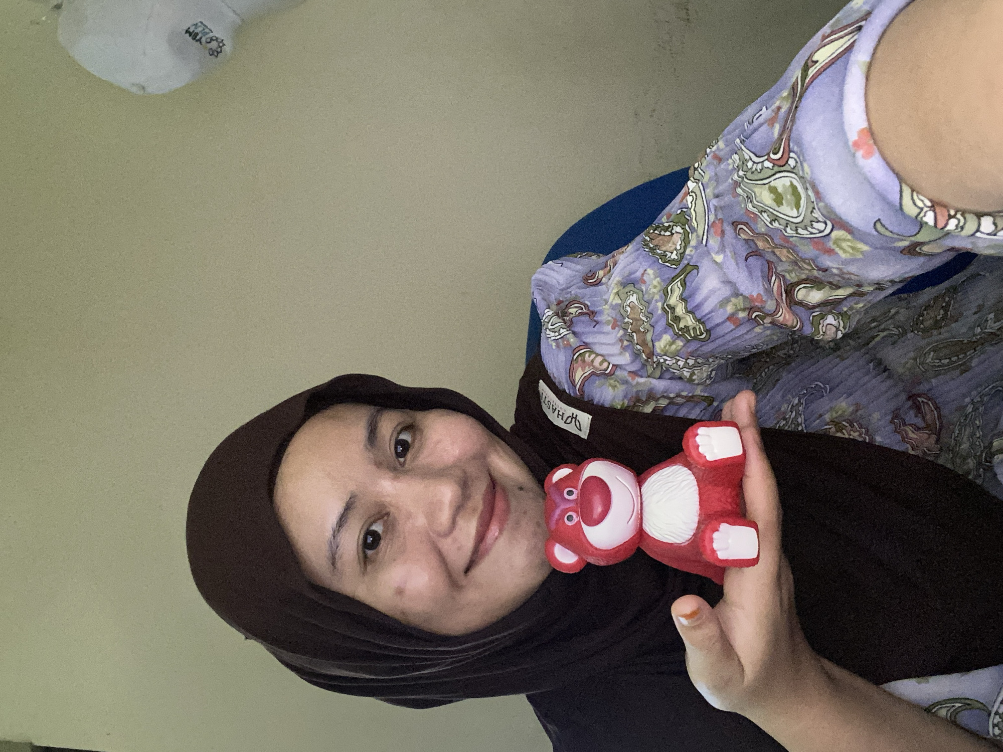 A woman wearing a dark brown hijab and a purple patterned top is holding a small red bear toy with white accents. She is smiling and looking at the camera, with her right hand visible holding the toy. The background is a light beige wall. To the left of the frame, a white object with some text is visible, possibly a light fixture. The setting appears to be indoors, with the woman sitting. The overall tone of the image is cheerful and casual. The visible text on the white object reads "YLM". There is also a tag on her top that is partially visible.
