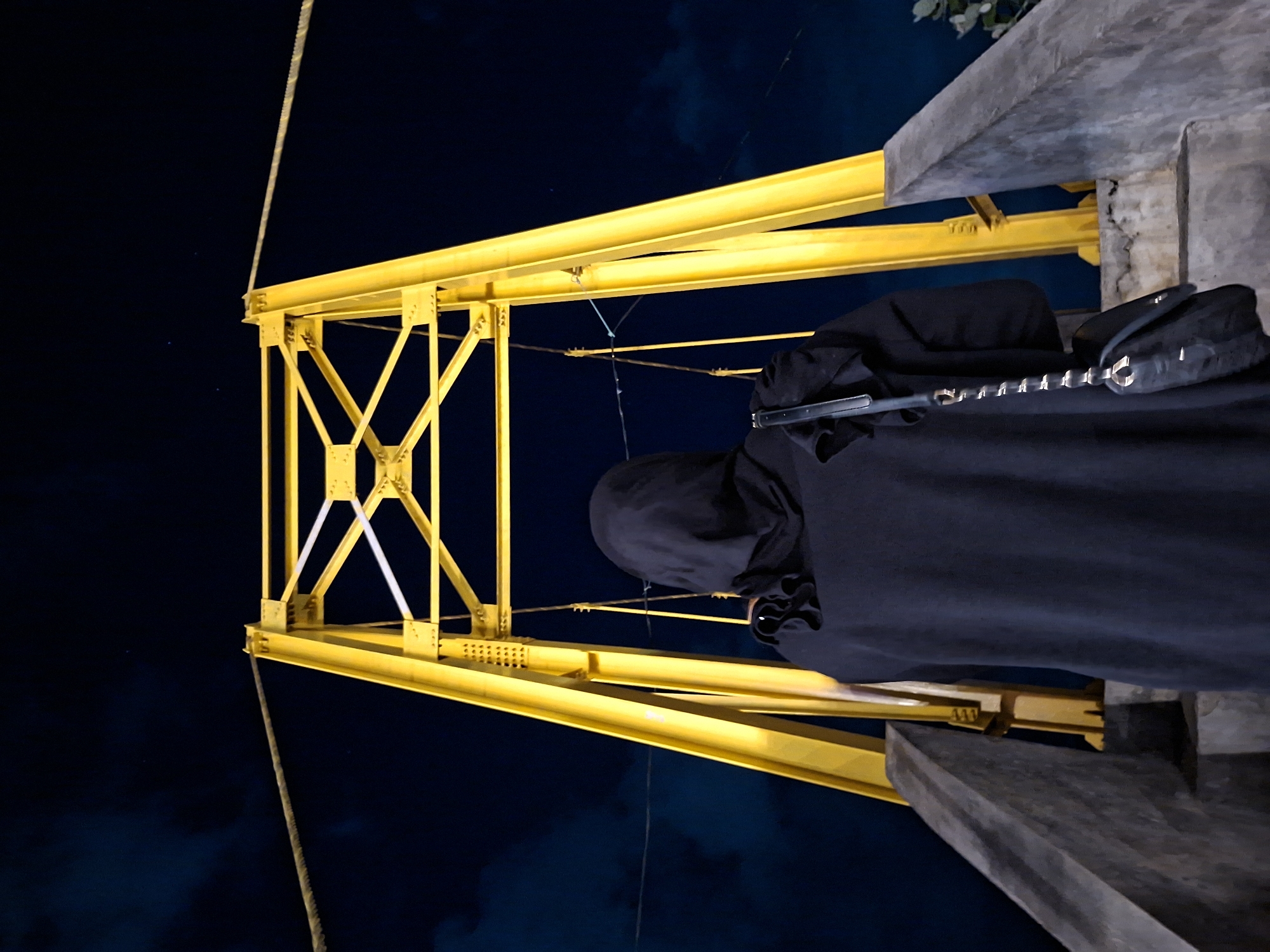 A person wearing a black head covering stands on a concrete structure in Lhokseumawe, Indonesia, next to a bright yellow steel bridge truss at night. The person faces away from the viewer and has a black purse with a decorative chain hanging on their side. A rope hangs from the truss into the dark blue sky. The setting is urban.