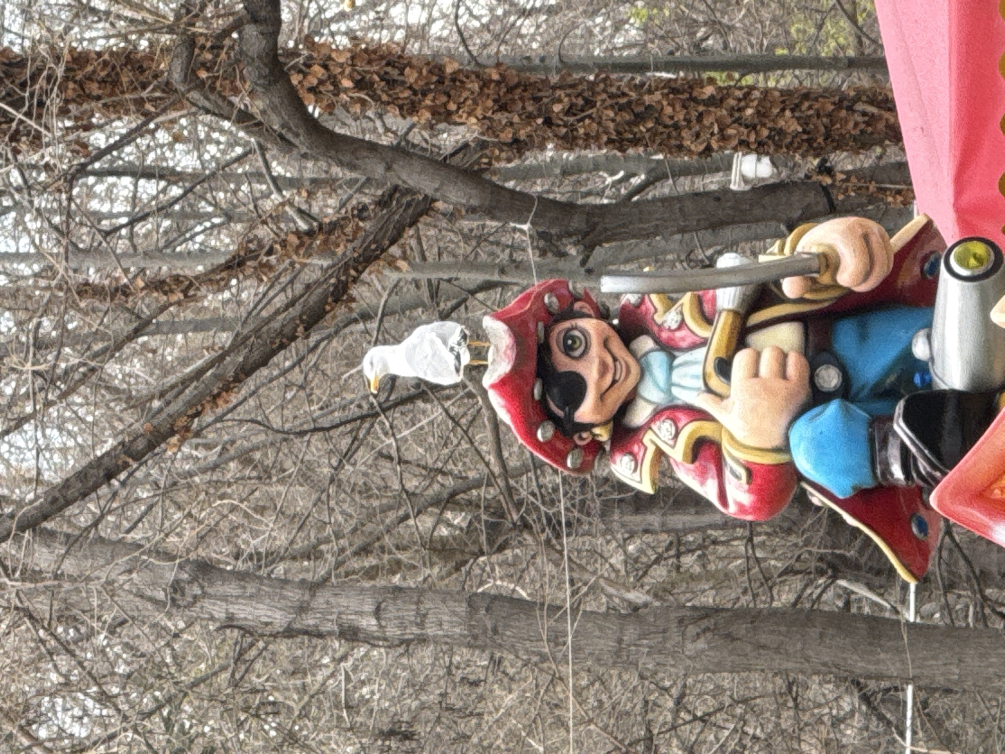An amusement park ride in Varna, Bulgaria, possibly a carousel or a swing ride, is observed with a pirate figure attached. The pirate is wearing a red hat, an eye patch, and a decorative red coat. A white bird, potentially a seagull, is attached to the pirate's head. The ride is set against a background of bare tree branches and some brown foliage, likely during the off-season or a colder time of year. A section of red canvas or fabric is also visible.