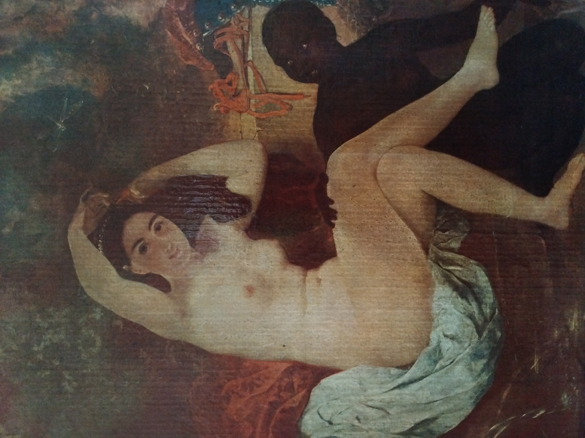 The painting depicts two figures in an indoor setting. On the left, a fair-skinned woman with dark hair is seated, facing forward and looking towards the viewer. She is unclothed, with her right arm raised and bent behind her head, and her left arm bent at the elbow, also behind her head. Her legs are crossed. She is draped with a light blue fabric on her left side. To the right of the woman, a dark-skinned individual is partially visible, crouching or kneeling, with their back to the viewer and their head turned towards the woman. They appear to be unclothed. The background consists of draped red fabric on the left, and a dark, indistinct area on the right with some decorative elements, possibly ropes or chains, in orange and blue. The overall environment suggests an opulent or theatrical setting. There is no visible connection to the city of Akhaltsikhe, Georgia.