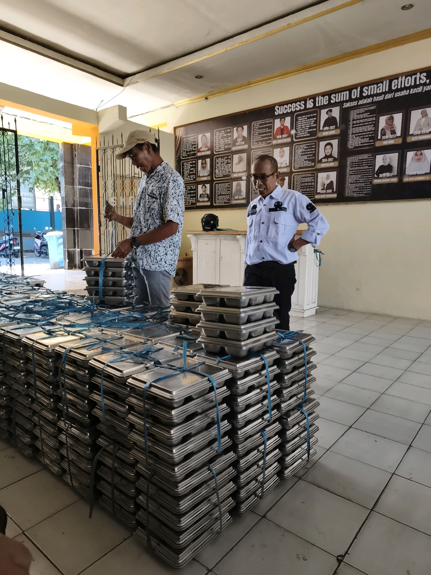 In an indoor setting, two men are present. The man on the left, wearing a patterned short-sleeved shirt and a baseball cap, is actively engaged in stacking metal trays. The man on the right, in a light blue collared shirt and dark pants, stands with his hands on his hips, observing. The trays are silver-colored and stacked in large quantities, secured with blue straps. The room features a tiled floor and a large bulletin board on the wall with photographs and text. The environment suggests an organized workspace, possibly a storage area. The overall scene is well-lit, with the presence of a doorway leading to an outdoor area visible in the background. The specific location in Makassar, Indonesia, is not explicitly indicated by any visible landmarks or signage within the image.