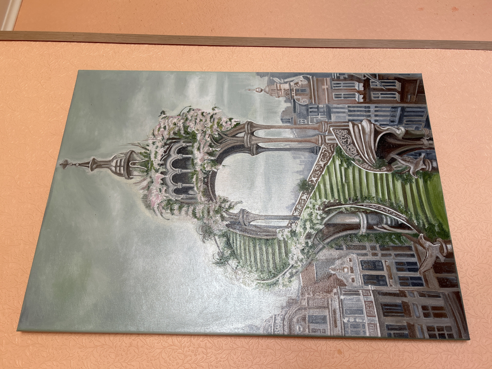 This is a painting of a fantastical spiral staircase adorned with flowers, ascending towards an ornate archway. The staircase is set against a backdrop of a European-style city with gabled buildings and spires. The painting is hung on an orange wall with a textured wallpaper pattern. There are no people or animals visible in the painting. The scene does not depict any specific location in Moscow, Russia, or any other real-world city.