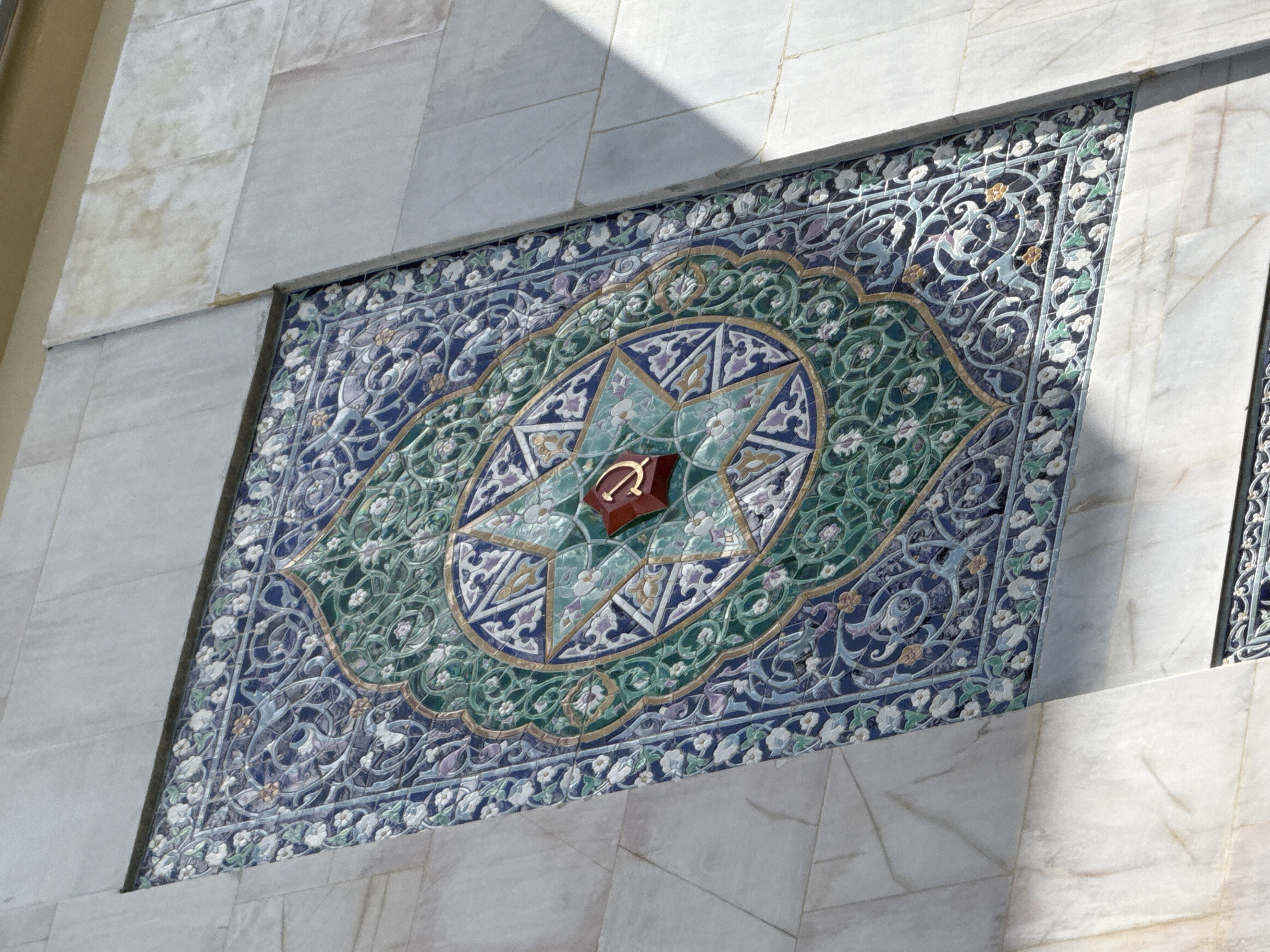This is a close-up, eye-level shot of an ornate tile mosaic adorning a building in Moscow, Russia. The mosaic is rectangular and features intricate geometric patterns and floral motifs in shades of blue, green, gold, and white. At the center of the mosaic is a stylized wooden object with a reddish-brown hue, possibly a religious or symbolic element.
The mosaic is set against a backdrop of light-colored marble tiles, which are arranged in a large grid. The marble is cool-toned and has subtle veining. A sharp shadow cuts across the top left portion of the marble, indicating bright sunlight and suggesting it is daytime, likely morning or afternoon. The overall impression is one of architectural beauty and detailed craftsmanship. There is no visible text in the image.