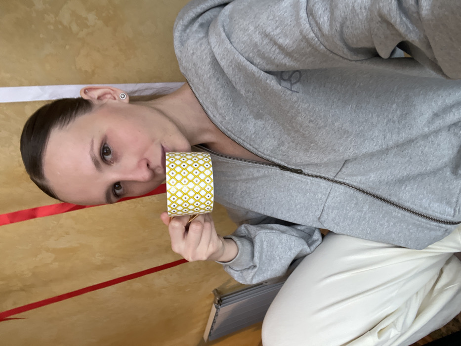 A female individual with light skin and dark hair pulled back is depicted in a medium shot, looking directly at the camera. She is holding a yellow and white geometrically patterned mug with a gold handle to her lips, appearing to consume a beverage. She wears a grey hooded sweatshirt with a visible zipper down the front and a dark 'RS' logo on the left chest, paired with light-colored, possibly off-white, trousers. A stud earring is visible on her right earlobe. The background consists of a textured, light brownish-yellow wall. Two red vertical strips are affixed to the wall on the left, and a single white vertical strip is visible on the right. A metallic-colored object, possibly a radiator, is partially visible in the lower-left background. The scene is located in Khimki, Russia.