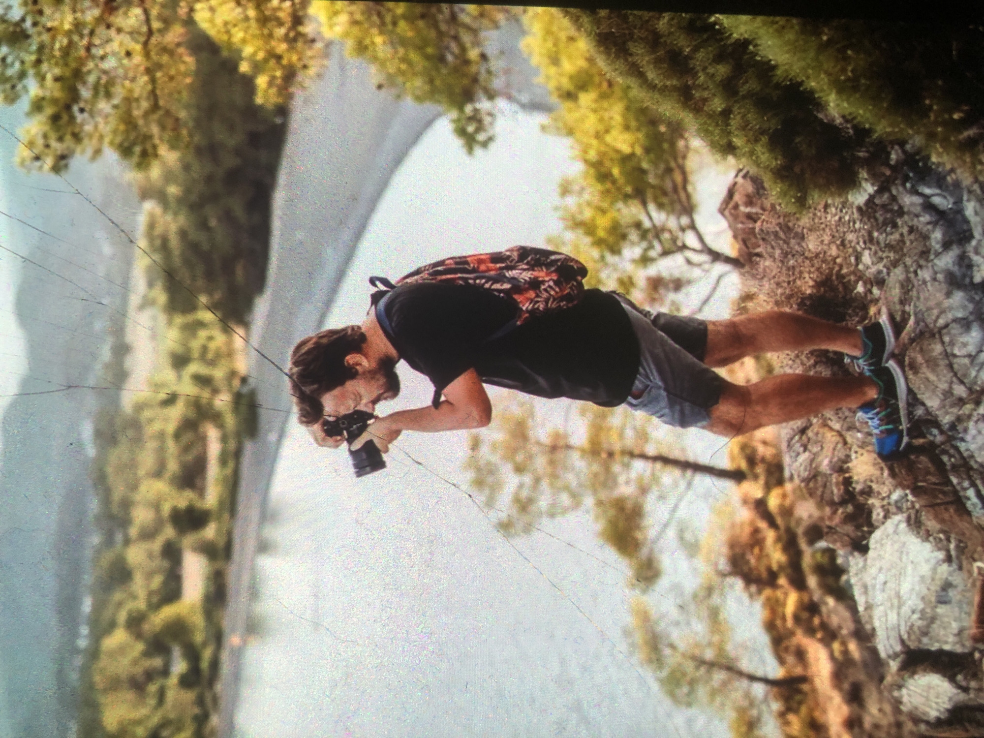 The image depicts a man with a beard, wearing a black t-shirt, grey shorts, and blue and black athletic shoes, standing on a rocky outcrop. He is holding a black camera to his face, seemingly taking a photograph. A backpack with a colorful pattern is visible on his back. The man is facing towards the left of the frame, looking out over a body of water, likely a bay or lake, which is bordered by a pebbled shore and lush green trees. The background features rolling hills covered in vegetation and a hazy sky. The scene appears to be a natural landscape, possibly a coastal or mountainous area. There is no indication that this location is in Tashkent, Uzbekistan; the environment suggests a different geographical region. The image has visible scratches and grain, indicative of its age or condition.