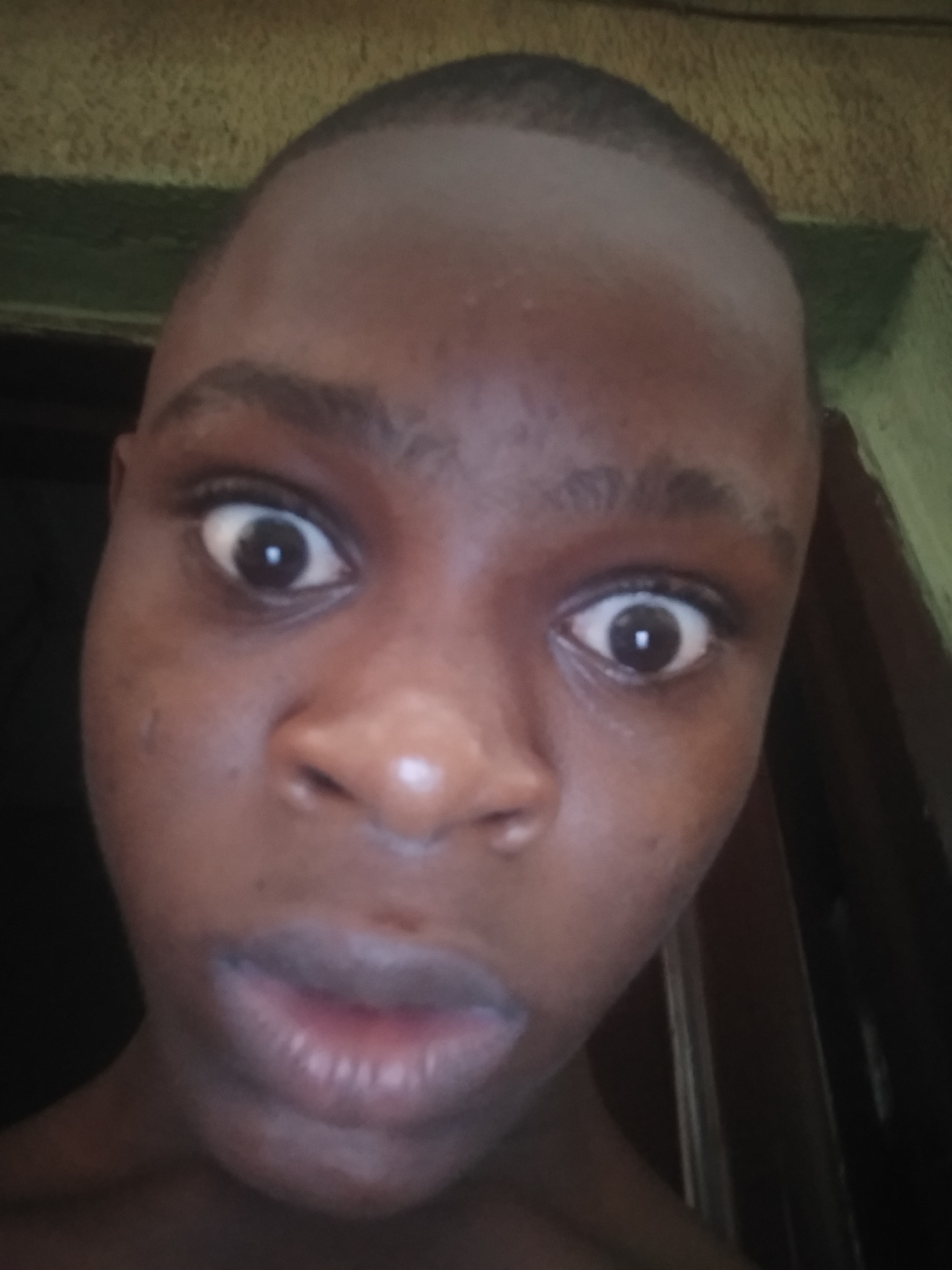 A close-up photograph shows a person with dark skin, short hair, and wide eyes. The person's mouth is slightly open. The background is indistinct, appearing to show a door frame. The location is unknown.