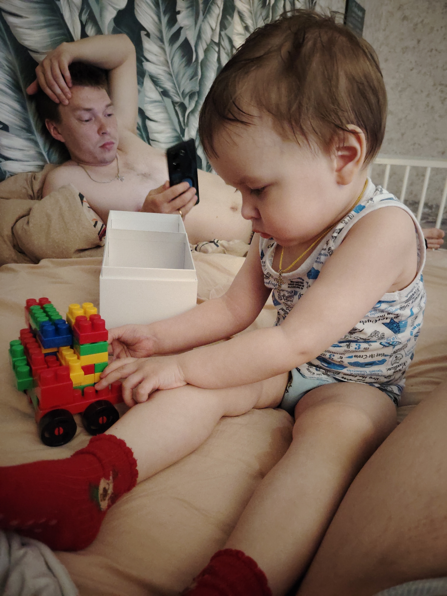 A young child is sitting on a beige bedspread, playing with colorful building blocks arranged like a vehicle. The child is wearing red socks, a white tank top with blue prints, and a small gold necklace. Next to the child is a white, open box. In the background, a man is lying in the bed against a headboard covered in a leafy pattern. He's shirtless and looking at a smartphone. The scene takes place inside a home, location Марковский, Russia.
