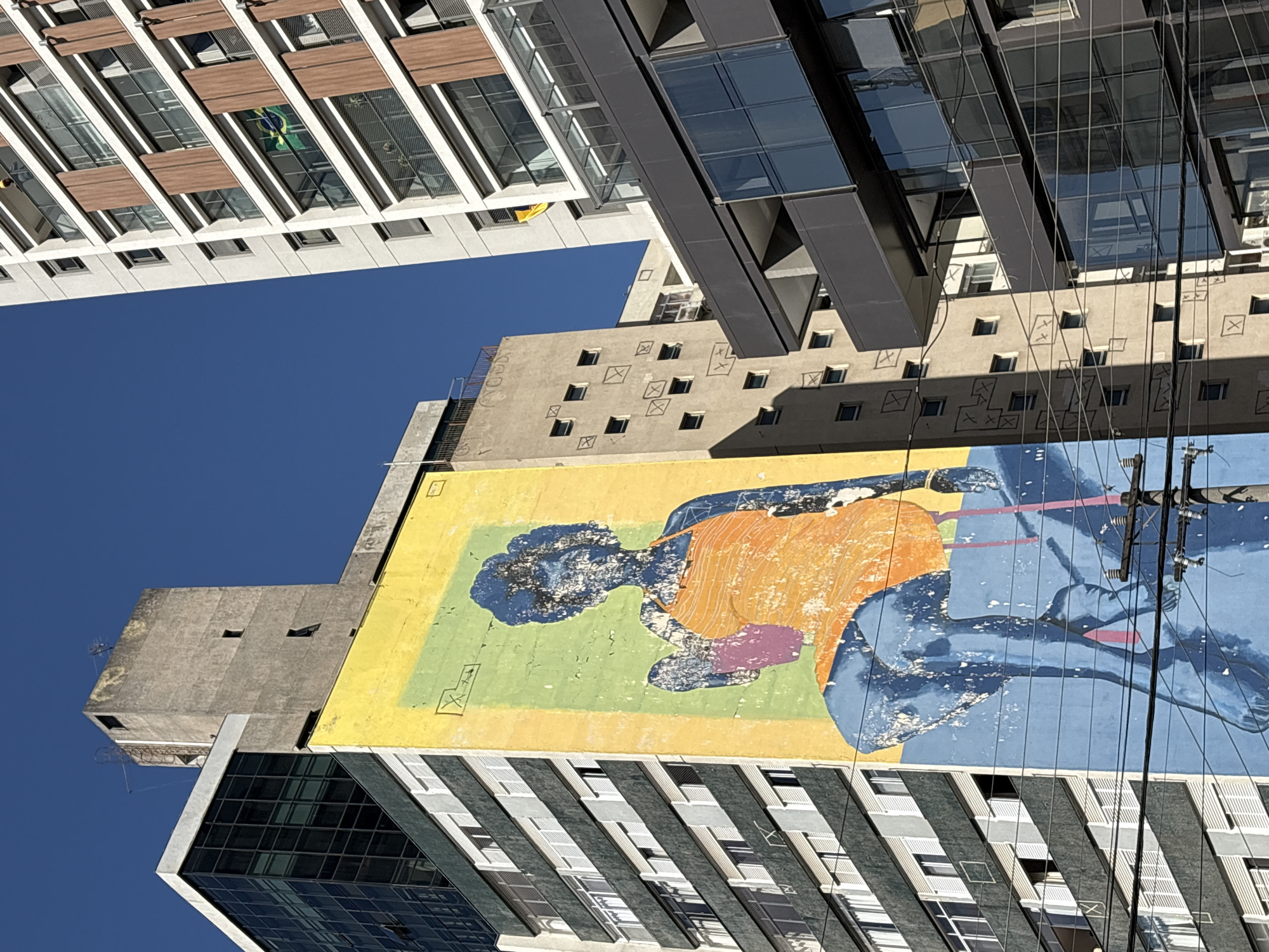 This media file captures a low-angle view of several modern buildings against a clear blue sky. The composition is dominated by the architecture, with one building featuring a large, colorful mural. The mural depicts a stylized human figure in shades of blue, orange, and pink, set against a yellow and green background. The figure appears to be in a relaxed, reclining pose.
The buildings themselves showcase varied architectural styles. The uppermost building has a white facade with horizontal wooden accents and rows of windows. To its right, a darker building with a glass facade juts out. Below these, a concrete building displays a grid of small, square windows, some of which have simple cross-like markings within them. The mural is painted on the side of a yellow-colored structure that seems to be integrated with or adjacent to the concrete building.
The perspective suggests the photo was taken from a lower vantage point, looking upwards. Power lines crisscross the frame, adding a layer of urban detail. The clear sky and bright sunlight indicate a daytime setting, likely on a fair-weather day. While no specific text is readily visible on the buildings or mural, the overall scene conveys a sense of urban dynamism and artistic expression within a city environment. The location context is São Paulo, Brazil.