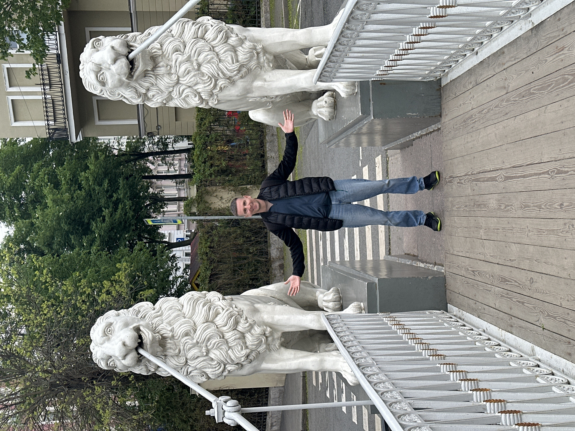 A full-body photograph shows a man standing on a bridge flanked by two white lion statues in Saint Petersburg, Russia. The man is wearing a black puffy jacket, a dark blue undershirt, blue jeans, and black tennis shoes with lime green accents. He has short dark hair and is smiling at the camera with both arms extended horizontally. The lion statues are positioned on gray pedestals at each end of the bridge. The lions have ornate, curly manes, and each holds a decorative white pole in its mouth. A white, ornamental railing lines the sides of the bridge. In the background, lush green trees and building facades of Saint Petersburg are visible. The bridge appears to be made of wooden planks.
