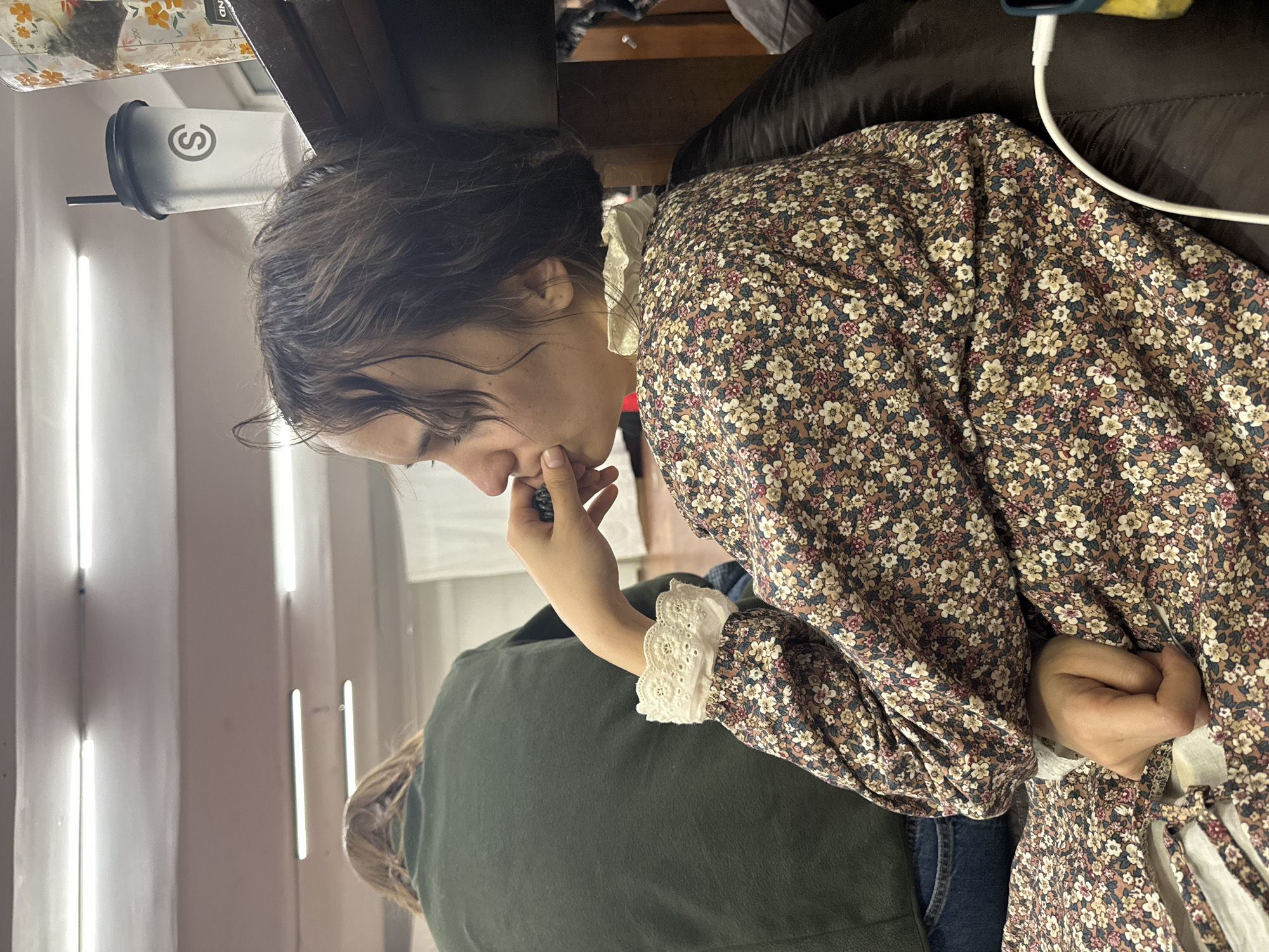 A woman is seated in an indoor location. The woman has dark hair and is wearing a floral dress with lace detailing on the sleeves and collar. She is holding an object near her face. A green jacket is visible on someone near her. A water bottle with the letter "S" on it is in the background next to a window. The location in the city of Moscow, Russia is unknown.