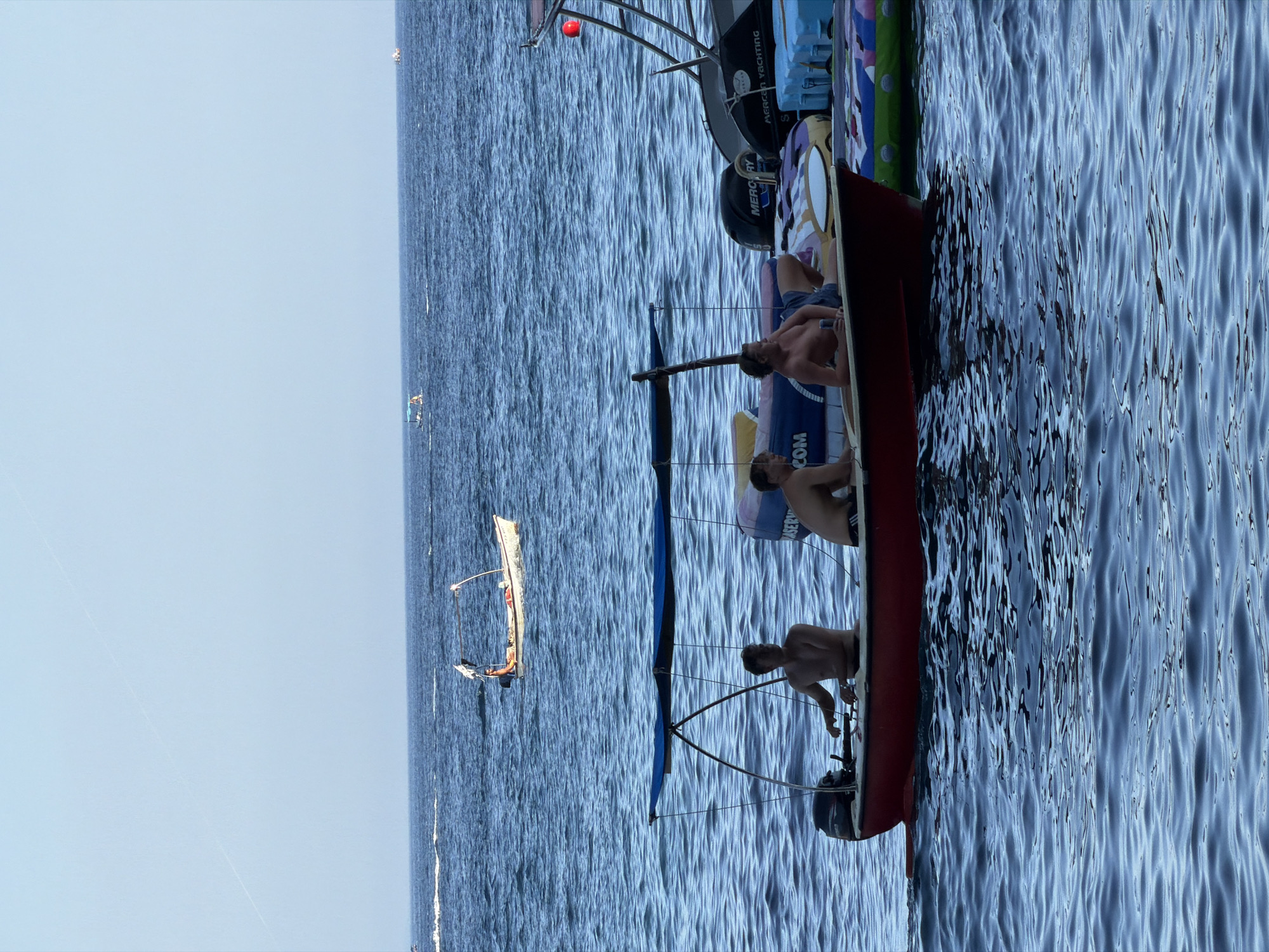 In Budva, Montenegro, on a clear day with a calm blue sea, three shirtless men are seated in a small red boat with a blue canopy. One man is at the helm, steering, while another reclines with a can in his hand, and the third sits upright, looking to the side. The boat is equipped with an outboard motor. In the background, other boats are visible on the water, and further out, the faint line of contrails stretches across the pale blue sky. The scene suggests a leisurely day spent on the water.