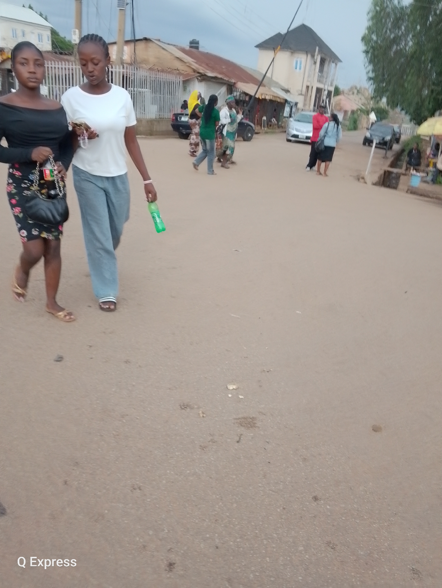 Two young women are walking on a dirt road in what appears to be a residential or market area. The woman on the left is wearing a black off-the-shoulder top and a floral print skirt. She is carrying a black handbag. The woman on the right is wearing a white t-shirt and gray pants. She is holding a green bottle of soda.
In the background, there are several buildings, some with corrugated metal roofs. There are also a few vehicles, including cars and what looks like a motorcycle or tricycle. Several other people are visible in the background, some walking and some standing. One person is wearing a bright yellow headscarf.
The sky is overcast, suggesting it may be late afternoon or early evening, and the weather appears to be calm. The overall scene depicts a typical day in a bustling community. The presence of shops and market stalls in the background suggests a commercial activity. The women appear to be casually walking and talking, possibly after shopping or on their way somewhere.
Visible text includes "Q Express" in the bottom left corner of the image, likely a watermark or branding. The setting appears to be a street in Jos, Nigeria, given the context provided.