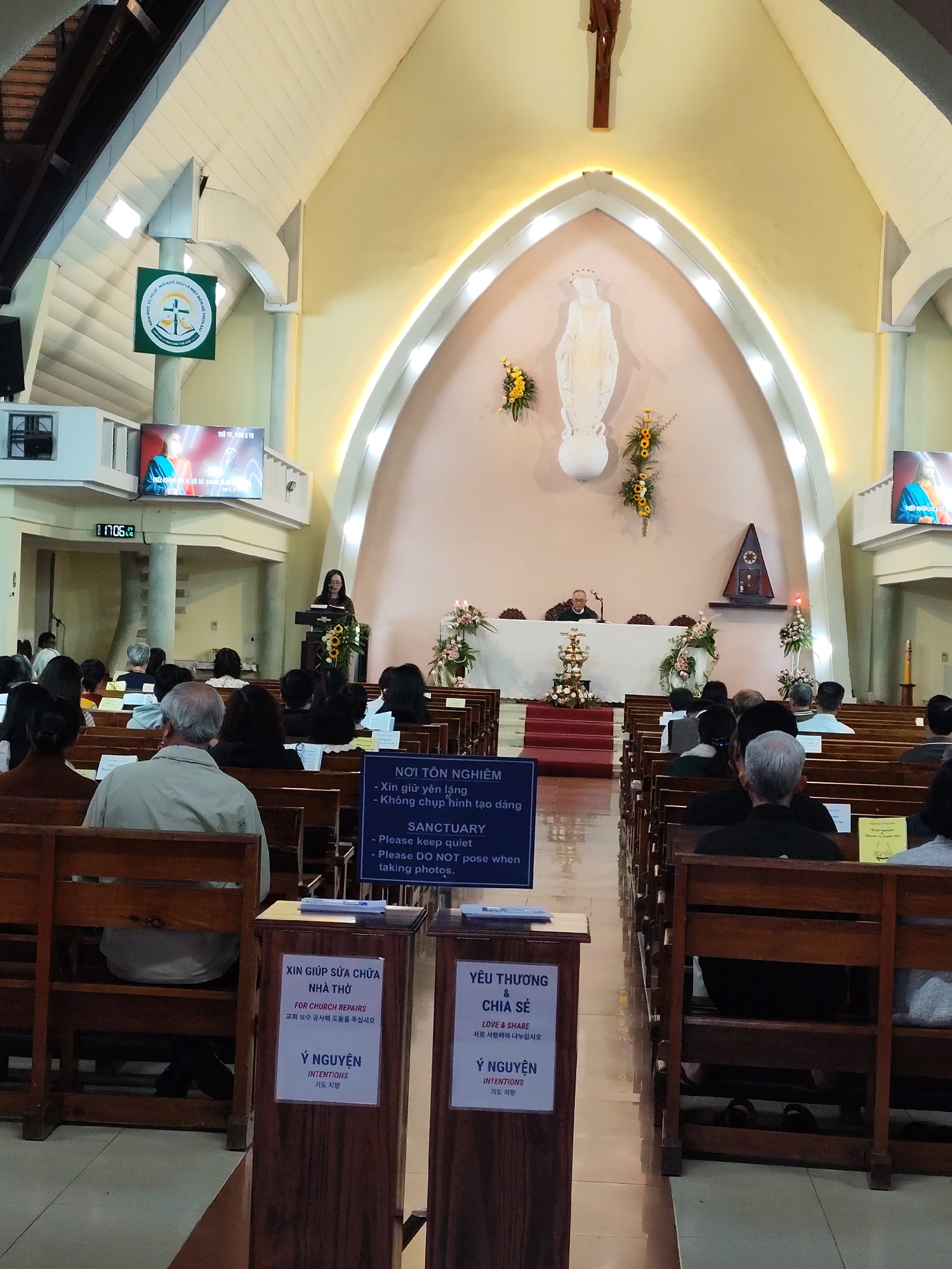 This media file depicts the interior of a church, likely in Vietnam, given the Vietnamese text visible. The scene is set during what appears to be a service or gathering, with many people seated in wooden pews facing a raised altar area. The church architecture features arched ceilings with soft lighting, and a statue of Mary is prominently displayed above the altar, flanked by floral arrangements.
In the foreground, two wooden donation boxes are visible, each with signage in Vietnamese and English. One box has text related to "FOR CHURCH REPAIRS" and "INTENTIONS," while the other is for "LOVE & SHARE" and also mentions "INTENTIONS." A sign on a pew in the middle ground indicates "SANCTUARY" rules, instructing visitors to "Please keep quiet" and "Please DO NOT pose when taking photos."
Several individuals are present: a woman stands at a lectern on the right side of the altar, appearing to address the congregation. A man is seated behind the altar, possibly a member of the clergy. The congregation members are diverse in age and are mostly facing forward, suggesting active participation or observation of the service.
Above the congregation, a digital clock displays "11:06," indicating the time of day. There are also screens mounted on the wall displaying religious imagery and text. The overall atmosphere seems reverent and orderly. The location cue of "Cam Ly - Đà Lạt, Vietnam" suggests this is likely Cam Ly Church in Đà Lạt.