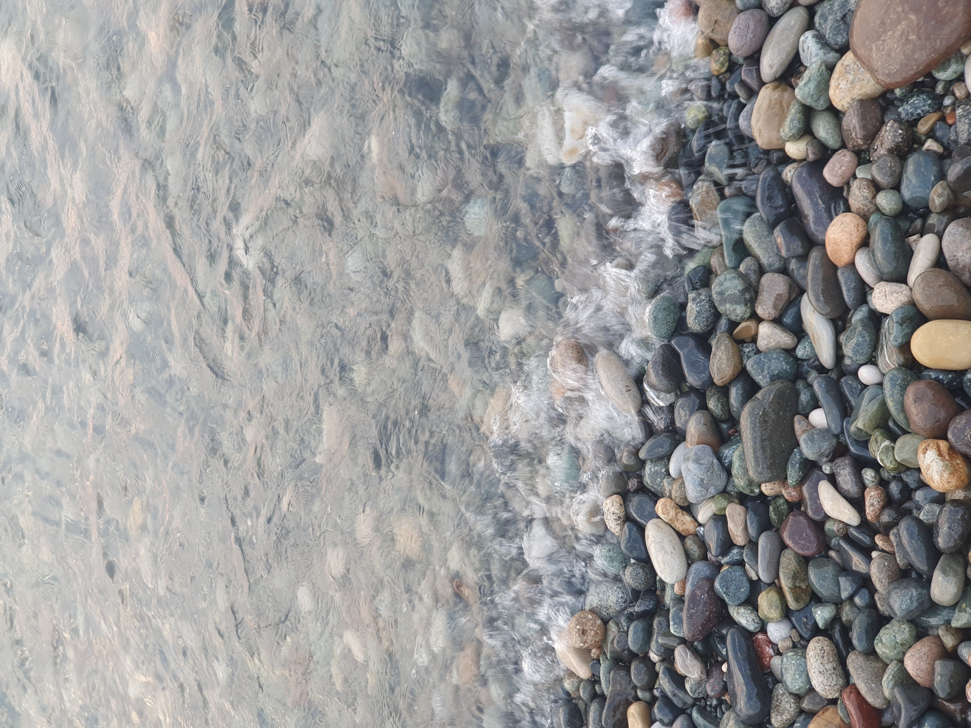 The image depicts a close-up view of a pebble beach with shallow, clear water gently lapping at the shore. The pebbles are of various sizes, shapes, and colors, ranging from dark grays and blacks to lighter browns, greens, and whites. Some of the pebbles are smooth and rounded, while others are more irregular. The water is slightly agitated, creating a foamy effect as it washes over the stones. The background is filled with more water, showing the reflections of the sky and the submerged pebbles beneath the surface. The scene is natural and serene, with no human presence visible. The lighting suggests it might be early morning or late afternoon, casting a soft glow over the scene. This location is consistent with the pebble beaches found along the coast of Batumi, Georgia, known for their natural beauty and the distinct pebble composition of their shorelines.