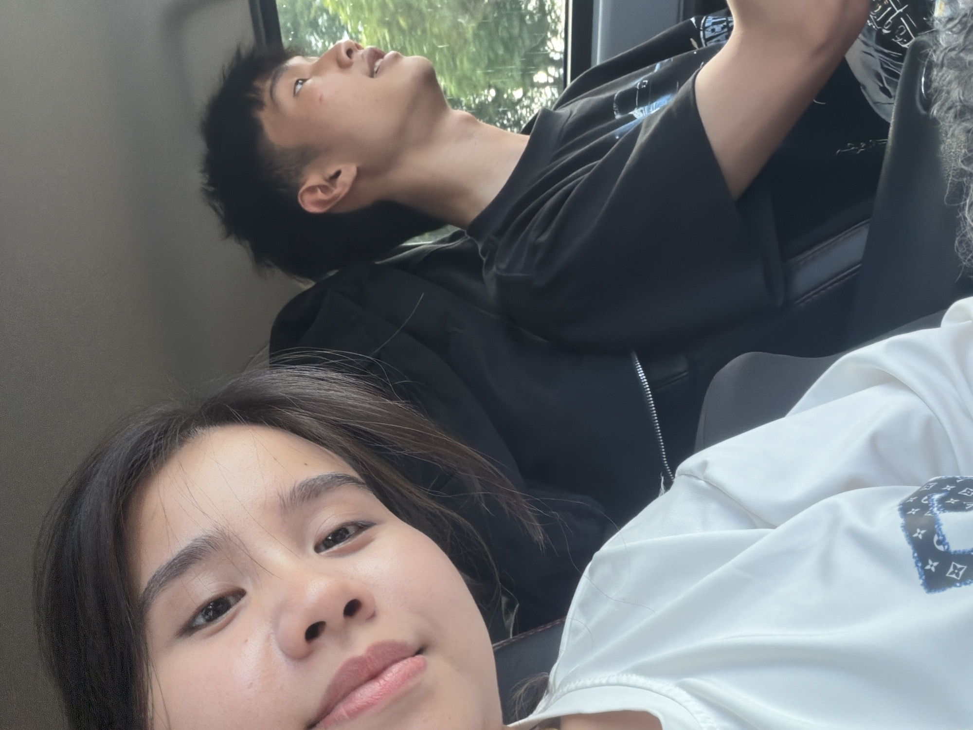 This image captures two individuals inside what appears to be a vehicle, likely a car, in Măng Đen, Vietnam. The shot is taken from a low angle, focusing on the person in the foreground and including the upper body of another person behind them.
In the foreground, a young woman is visible, taking a selfie or being photographed from close range. She has dark hair, a clear complexion, and is looking directly towards the camera with a neutral to slightly pleasant expression. She is wearing a white top, on which a blue and black pattern resembling the Louis Vuitton monogram is visible.
Behind her, positioned higher and further back, is a young man. He has short, dark hair and is looking upwards and out of a window, which shows glimpses of green foliage outside, suggesting trees or bushes. He is wearing a black long-sleeved top or jacket. His Adam's apple is noticeable. His right arm is bent, and he appears to be holding a phone or small object, though its details are obscured. On his black shirt, some white, indistinct text or design is present, possibly Arabic script, but it is not clearly legible.
The lighting is bright, indicating it is daytime. The people appear to be either resting or traveling, with the foreground person engaging directly with the camera and the background person looking away, perhaps at the passing scenery. The setting suggests a casual moment during travel.