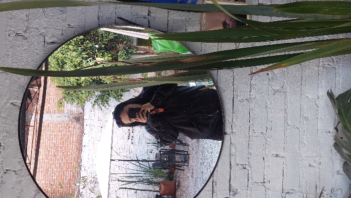 A circular, black-framed mirror, mounted on a white-painted, distressed brick wall, captures a reflection selfie within an outdoor commercial establishment in Tlaxcala City, Mexico. The reflection shows an individual, presumed female, with dark hair, wearing a dark zippered hoodie and sunglasses pushed onto her head. She holds a dark smartphone, featuring a visible orange charm, obscuring her face as she photographs her reflection. The environment reflected in the mirror includes an outdoor patio area with multiple dark metal tables and chairs positioned on a light-colored ground surface. A white umbrella or canopy provides shade. Various potted plants are arranged around the patio. In the upper background of the reflection, a red brick wall is visible, along with a dense green tree. A partially mirrored and inverted sign, possibly reading "WELCOME," is discernible. To the right of the mirror, outside the reflection, tall, sword-like green plants are prominent, and a large green leaf is visible at the bottom of the frame.