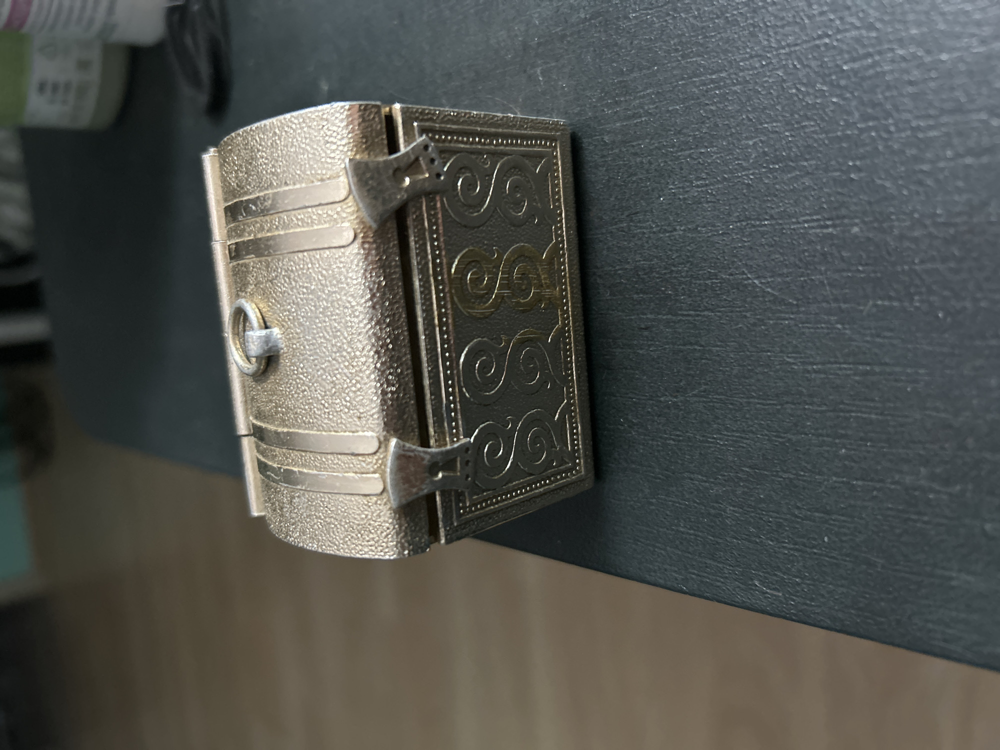 A close-up, slightly angled shot shows a small, ornate treasure chest-shaped object on a dark, textured surface. The object is metallic and gold-colored, with a textured finish on the lid and intricate decorative patterns on the front. The lid has a ring at the top and two decorative clasps on the front. The front of the chest features a design with multiple "S" shapes and decorative borders. The background is blurred, showing a light-colored floor and some indistinct objects. The location is not discernible from the image, and there is no indication of Tugulym, Russia.