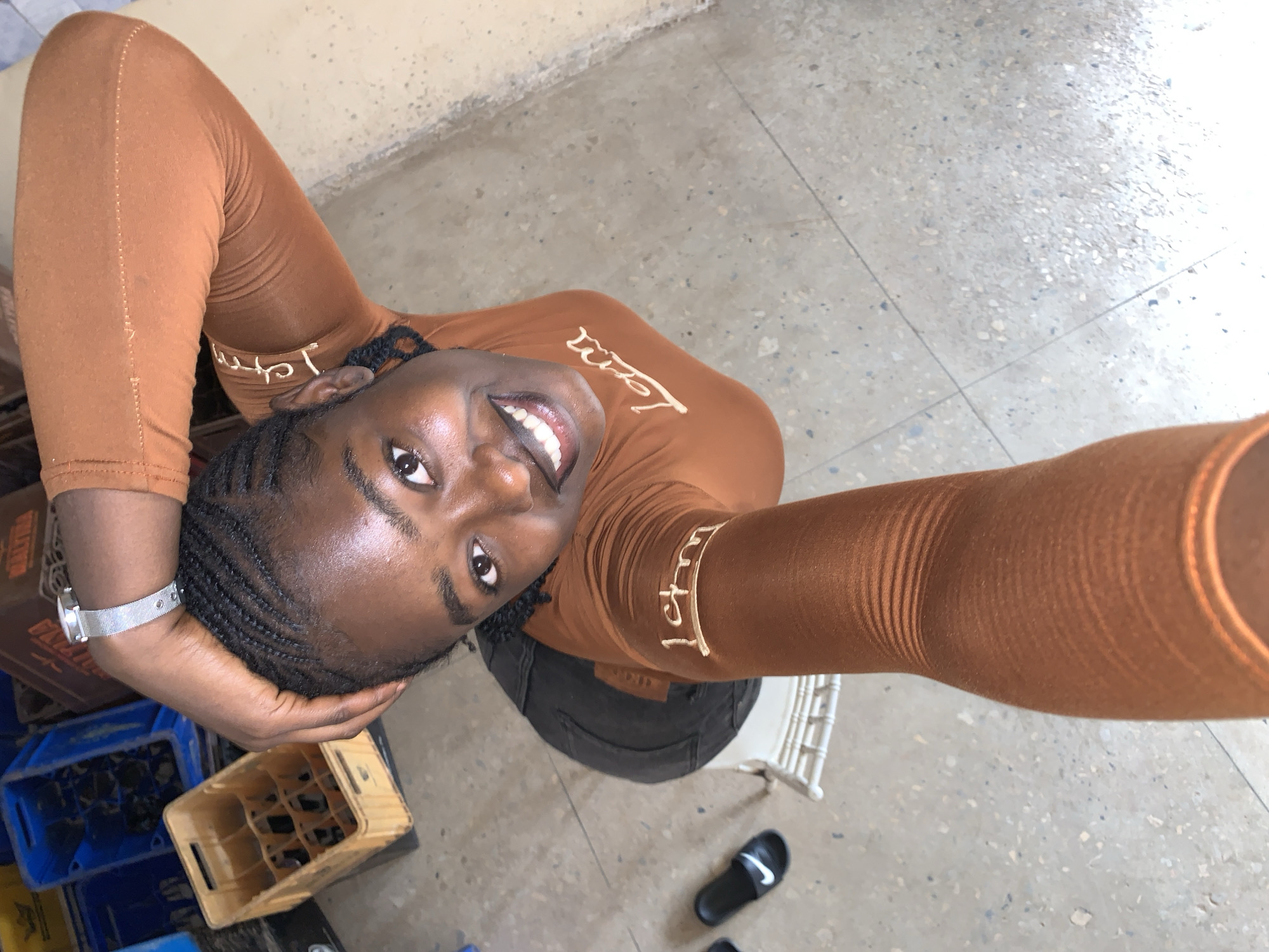 A high-angle selfie captured in the city of Jos, Nigeria, features a young woman with dark skin and braided hair (cornrows), smiling directly at the camera. She is wearing a fitted, long-sleeved, rust-colored top with light-colored lettering; "1cm" is visible on her left sleeve, and "I am" or "I'am" is discernible on the upper chest. A silver-toned wristwatch with a mesh band is present on her left wrist. Her left hand is positioned atop her head. The background on the left side of the frame includes multiple stacked plastic beverage crates, some blue and one light-colored with empty compartments. On the lower left, a black slide sandal with a white Nike swoosh logo is visible, alongside a partial white chair frame. The floor is a light-colored, speckled terrazzo or concrete surface with visible grout lines.