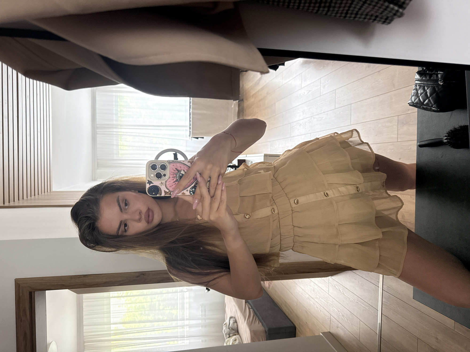 An indoor mirror selfie captures a young woman standing in what appears to be a modern residential space in Plovdiv, Bulgaria. The image is rotated 90 degrees counter-clockwise. The woman, with long, dark brown hair and a neutral expression, looks directly into the camera. She holds a smartphone with a distinctive pink case featuring a design that appears to be an abstract eye or lips, adorned with sparkling elements around its three camera lenses. Her nails are painted light pink, and a thin red string bracelet is visible on her left wrist.
She is dressed in a light beige or tan short dress or romper with gold-colored buttons down the front and multiple ruffled layers on the skirt section. The reflection in the mirror reveals a room with light wooden floorboards and white walls. To the left, a decorative wooden slat wall is visible. In the background reflection, a window with horizontal blinds allows natural daylight to stream in, showing hints of greenery outside, suggesting it is daytime. A bed with a brown cover is also reflected behind her. To the right of the frame, a black quilted bag and a black hairbrush hang on a dark piece of furniture or stand. The overall setting appears to be contemporary and well-lit.