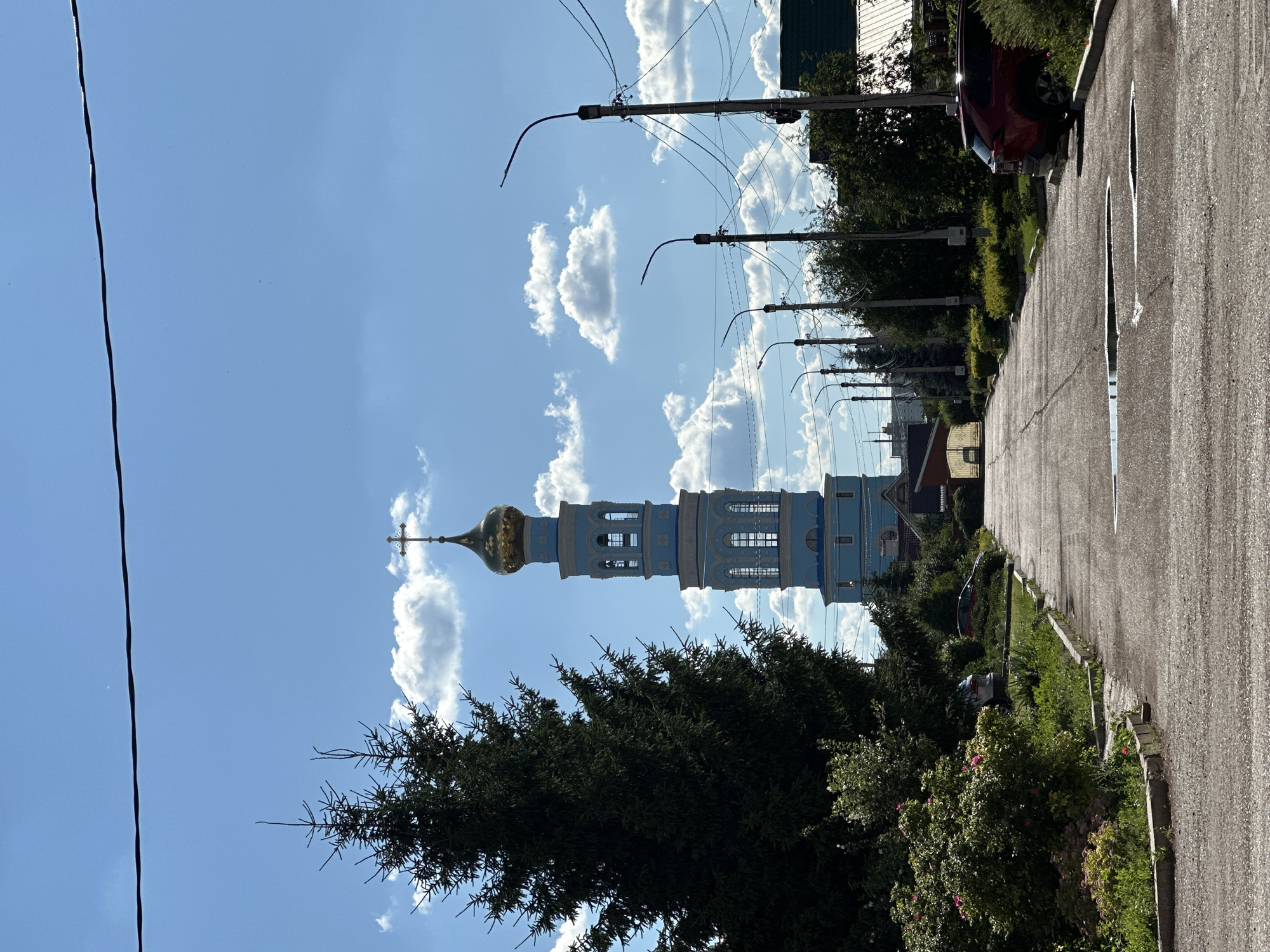 In the city of Mariinsky, Russia, a bright blue bell tower with a golden dome, topped by a cross, stands tall against a clear blue sky with scattered white clouds. The tower is flanked by a large, dark green coniferous tree on the left. A paved road leads towards the tower, with a row of streetlights on the right side of the road and lush greenery and bushes lining both sides. A red SUV is parked on the right side of the road, partially obscured by bushes. The road surface shows signs of wear and has several large puddles of water. In the background, beyond the streetlights, several houses with green roofs are visible.