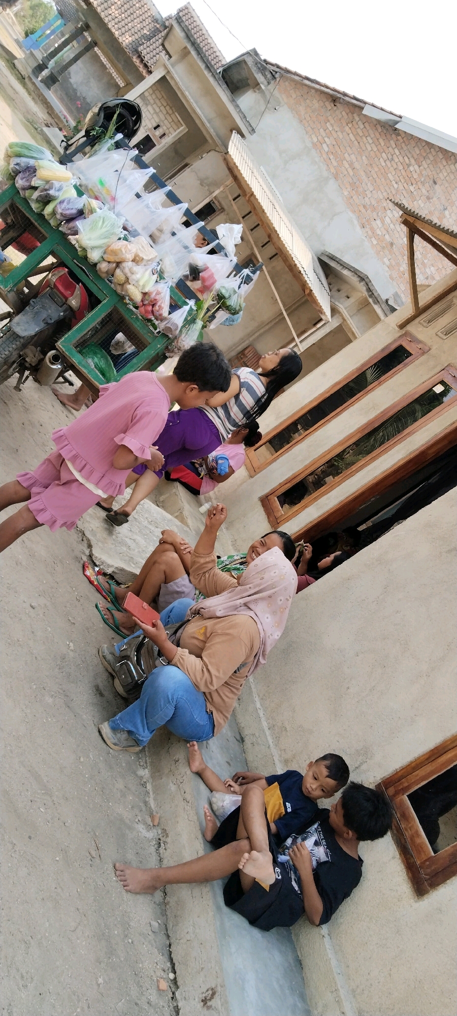 The image shows an outdoor scene in Tanjung Ratu Ilir, Indonesia, with several people and objects. In the foreground, three children are seated on the ground. Two boys, one wearing a dark blue and yellow shirt and black shorts, and the other in a black t-shirt with a graphic, are positioned close together, looking at each other. A third child, wearing a dark blue and yellow shirt, is sitting next to them. Behind them, a woman wearing a light brown long-sleeved shirt and blue jeans is seated, holding a red wallet and looking to her left with a smiling expression, her right arm raised. She is wearing a light pink headscarf. A young boy in a pink ruffled outfit is standing to the left of this woman, facing away from the camera and towards a cart.
The cart, loaded with various produce in plastic bags and some loose vegetables, is positioned in the background. A red and silver motorcycle is attached to the front of the cart. Behind the cart and the people, there are buildings with brick and concrete walls, tiled roofs, and wooden window frames. The ground is paved with concrete and appears somewhat cracked. The overall lighting suggests it is daytime, possibly late afternoon.