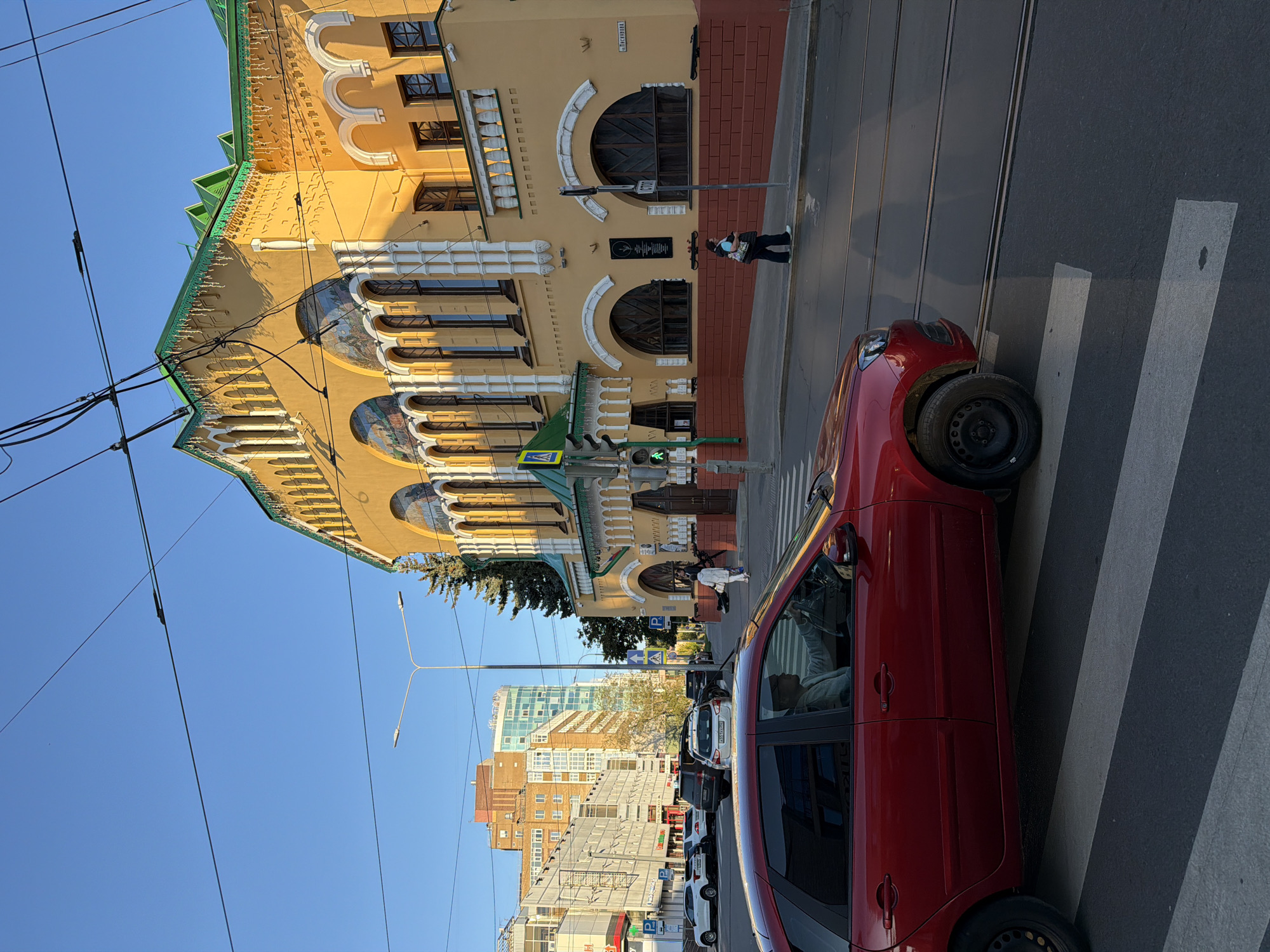 A bright red hatchback is stopped at a pedestrian crossing on a street in Nizhny Novgorod, Russia. The car is facing left, with the driver visible behind the wheel. To the right of the car, a yellow building with green trim and arched windows is prominent. A person walks along the sidewalk in front of the building. In the background, modern high-rise buildings and trees are visible under a clear blue sky. Overhead, electrical wires crisscross the frame. The scene appears to be in the late afternoon or early evening, judging by the lighting and shadows.
