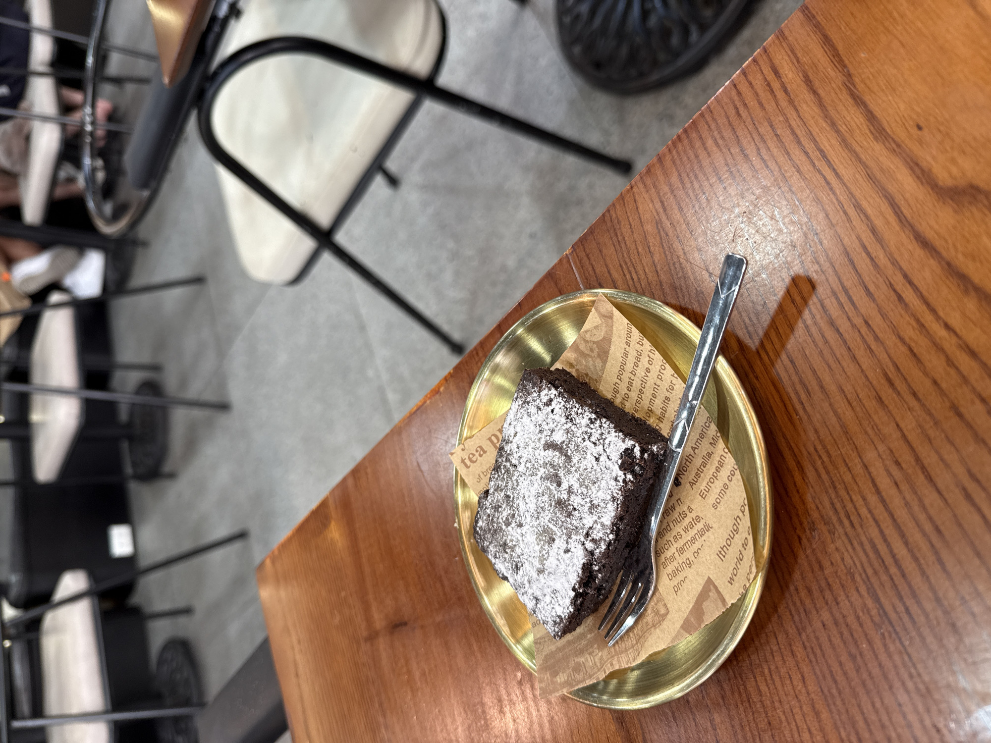 The image shows a brownie on a small gold plate with a fork, set on a wooden table. The brownie is covered with powdered sugar. The plate rests on a piece of paper with printed text. In the background, there are chairs and other furniture. There is no indication of the city of Đà Nẵng, Vietnam, or any people present in the immediate scene.