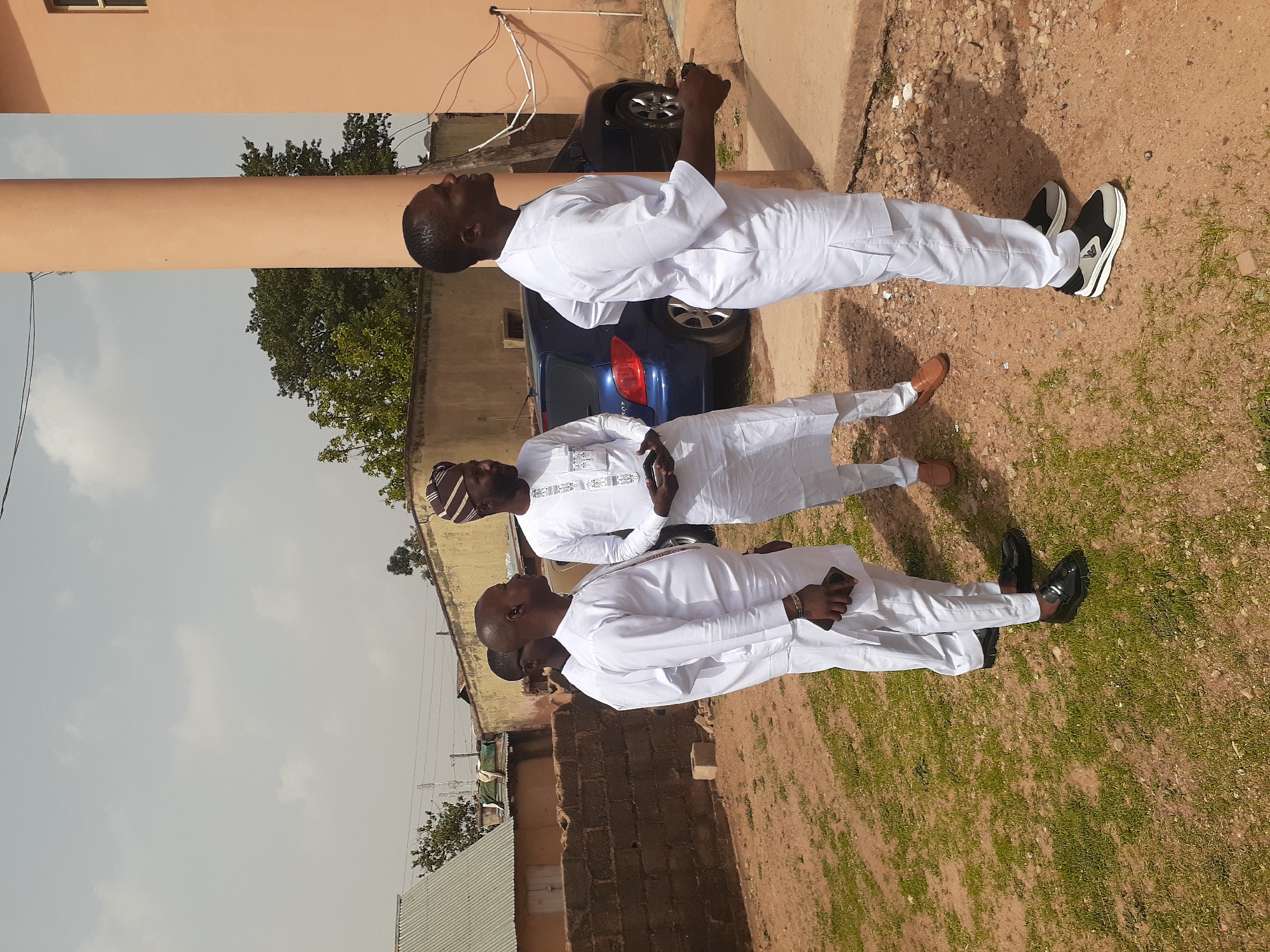 Three men are standing outdoors in what appears to be a residential area in Unknown, Unknown. All three men are wearing white outfits. The man on the right is wearing black and white sneakers, the man in the middle is wearing brown shoes, and the man on the left is wearing black shoes. A dark blue car is parked behind the men, near a peach-colored building with a dark window. Behind them are other buildings of brick and metal, with power lines visible. The ground is a mix of dirt and sparse grass.