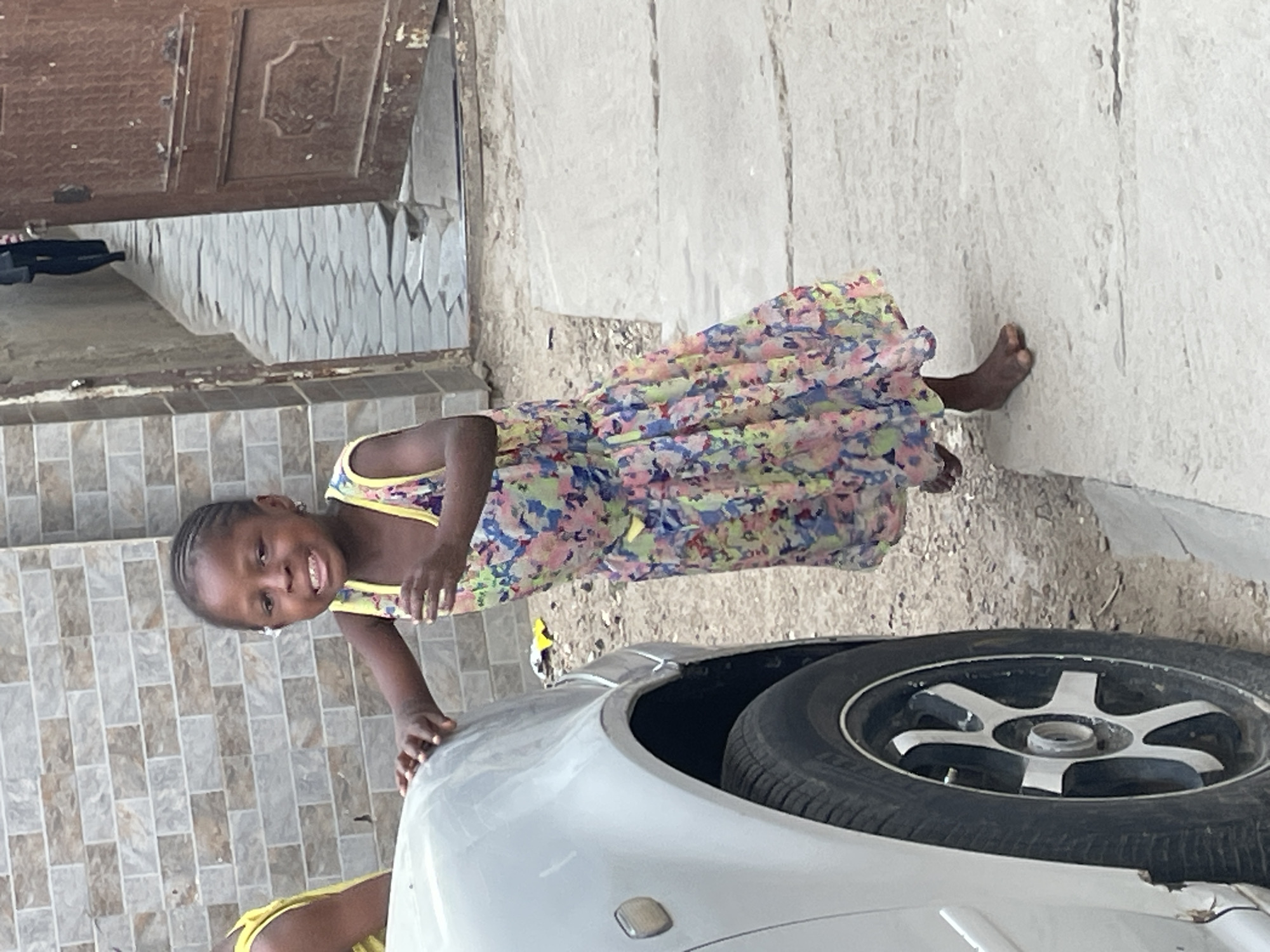 A young girl stands next to a light-colored car with a visible spare tire in the trunk. She is wearing a floral dress with yellow trim and smiling. The background shows a tiled wall and a textured, off-white exterior wall. The location appears to be a residential area in the city of Unknown, Unknown.