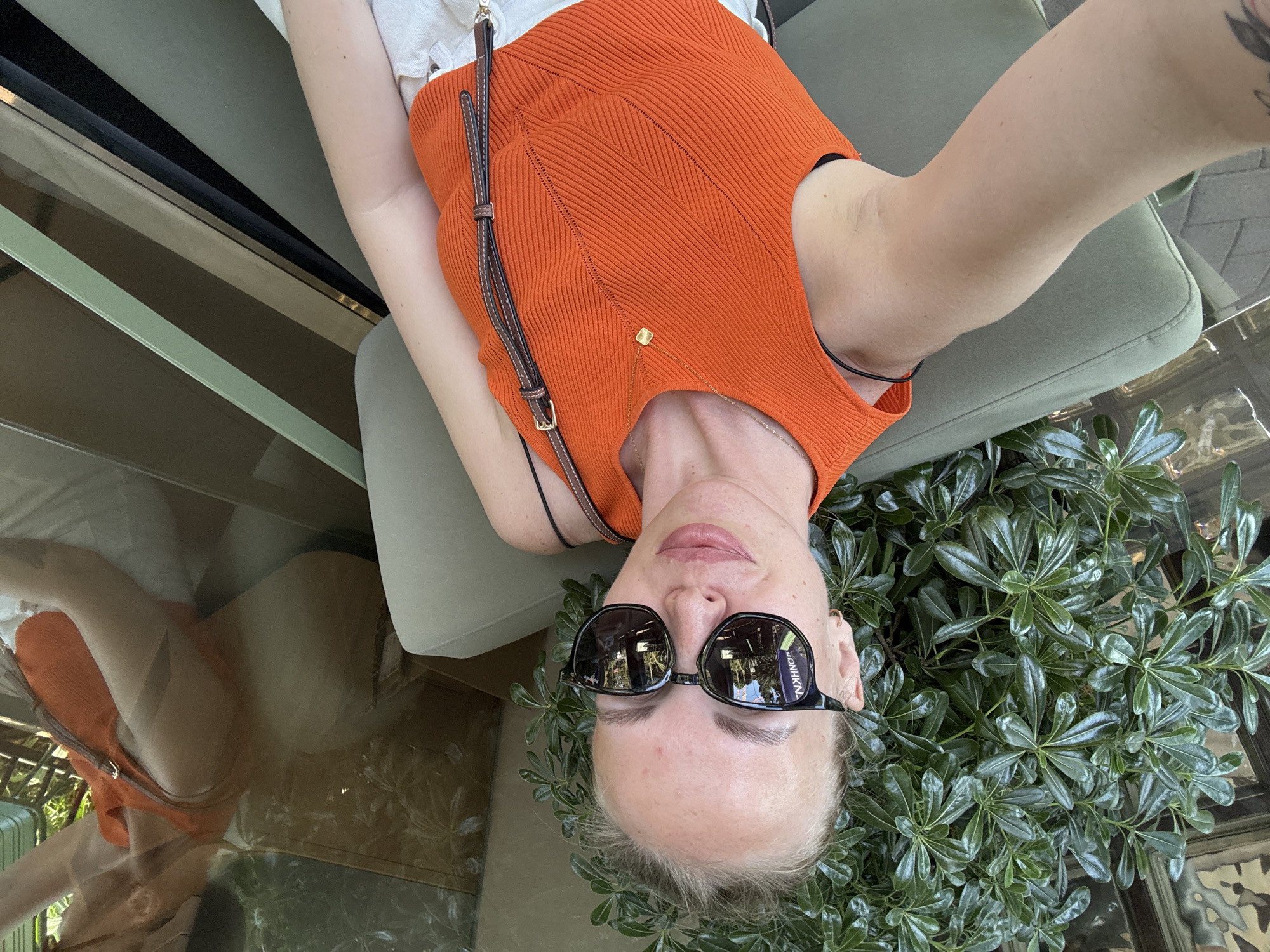 A woman with blonde hair pulled back, wearing sunglasses and an orange top, takes a selfie while seated outdoors. She is leaning against a muted green cushioned seat, with a lush green plant to her left. Her arms are bare, and she has a brown strap from a bag draped across her chest. The background includes reflections in what appears to be glass, showing more greenery and the environment of a cafe or public space. There is no information to indicate the location is Sochi, Russia.